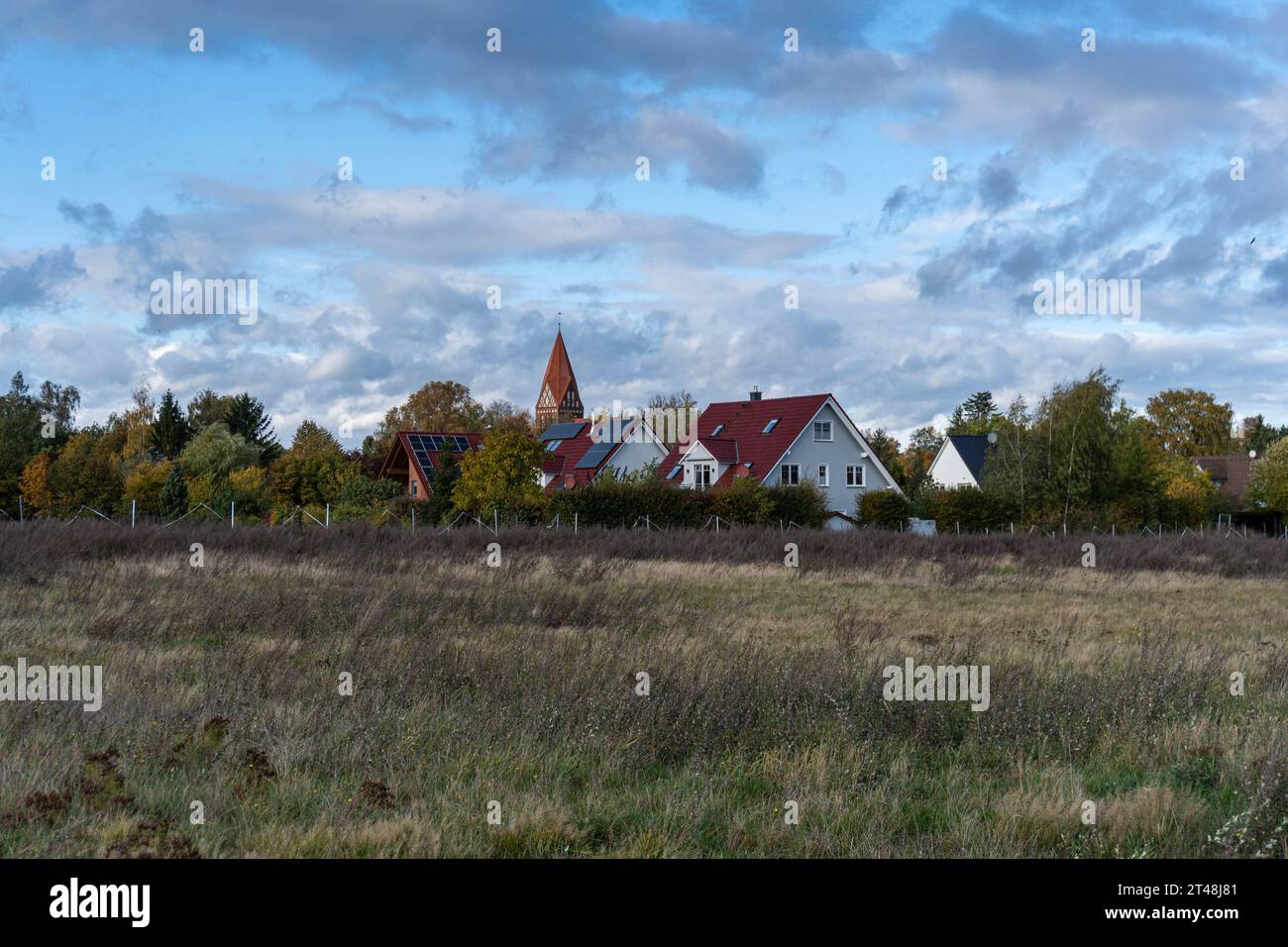 tranquillo villaggio con case dai tetti rossi e una lontana guglia della chiesa confina con un campo autunnale aperto sotto un cielo parzialmente nuvoloso, evocando un tranquillo fascino rurale Foto Stock