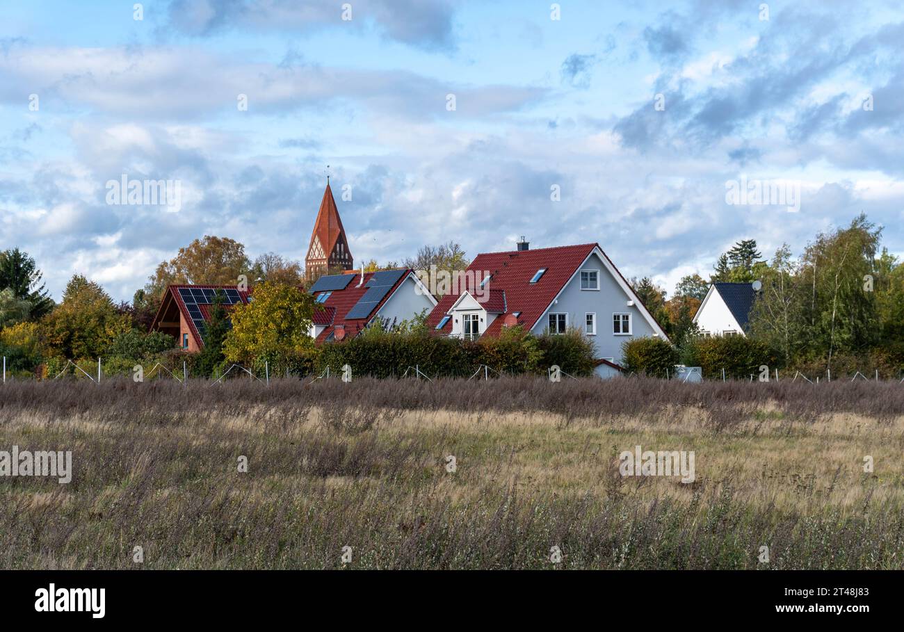 tranquillo villaggio con case dal tetto rosso e un campanile di chiesa sorge dietro un campo autunnale aperto sotto un cielo parzialmente nuvoloso Foto Stock