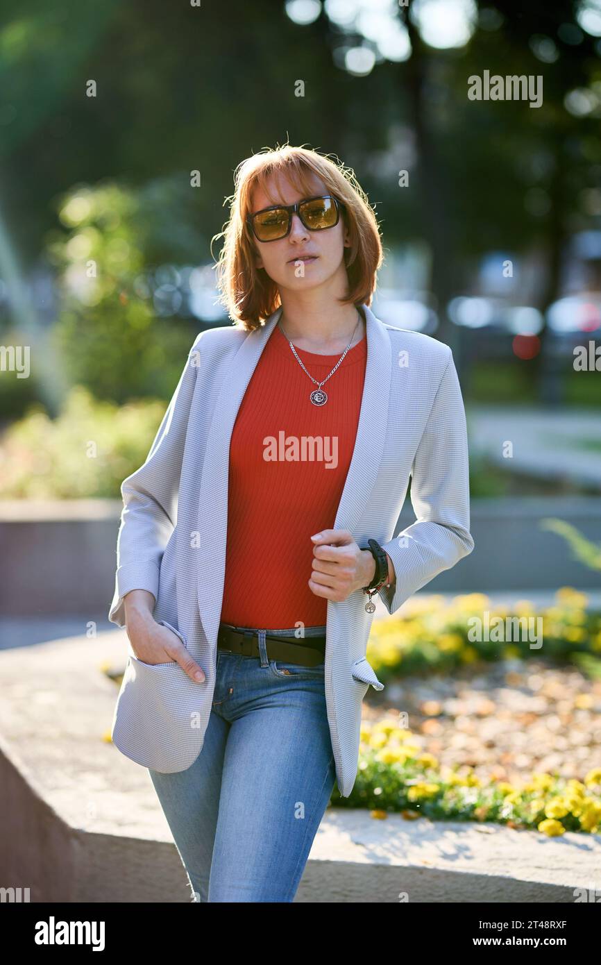 Una donna alla moda rossa che si gode una serata autunnale in città. Indossa una giacca bianca alla moda, una t-shirt rossa vivace e jeans jeans makin Foto Stock