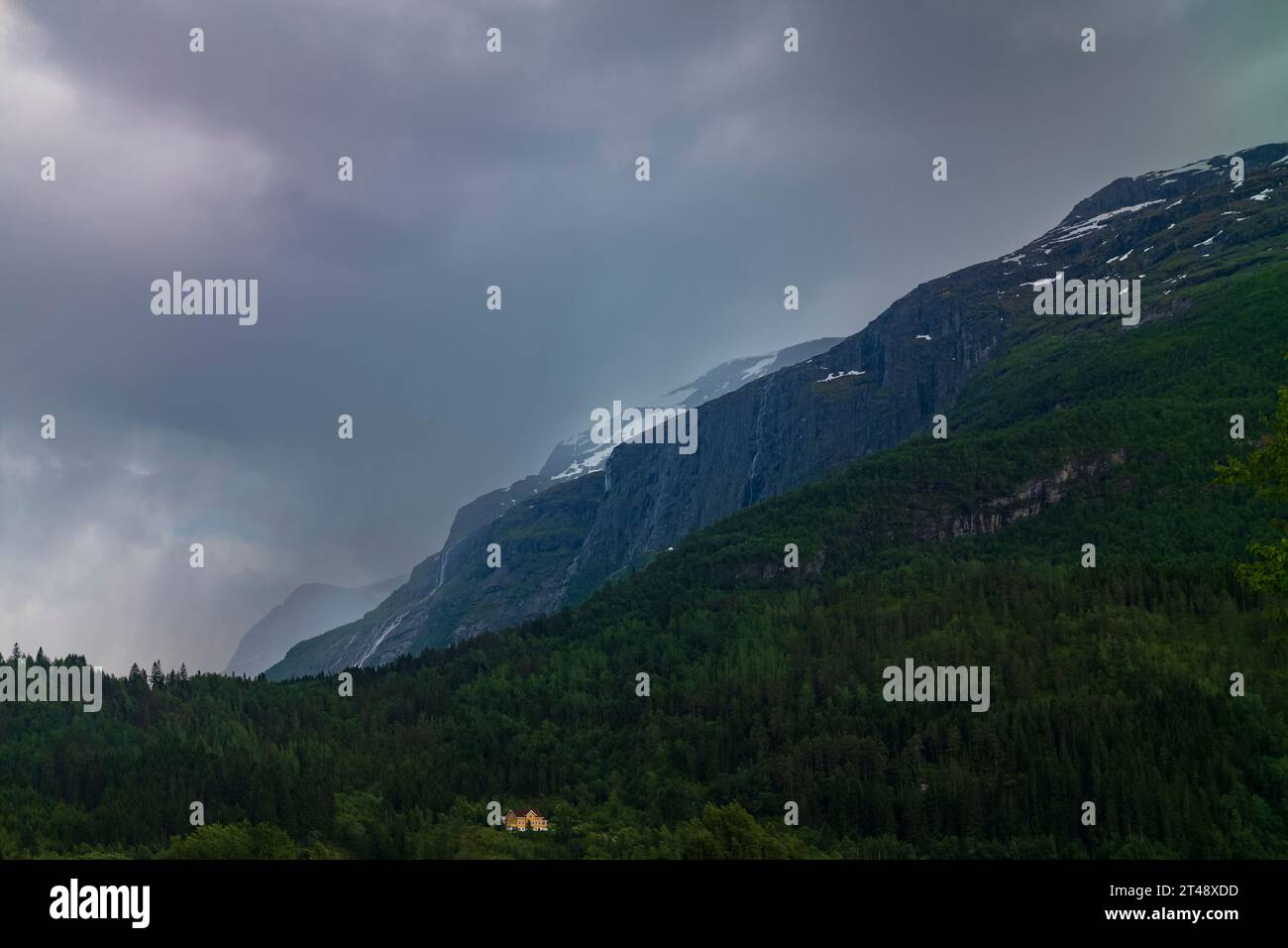 Una casa solitaria, gialla, poggia sul fianco di una collina ed è in contrasto con le verdi montagne sopra, a Loen, in Norvegia, durante una tempesta, la sera d'estate. Foto Stock