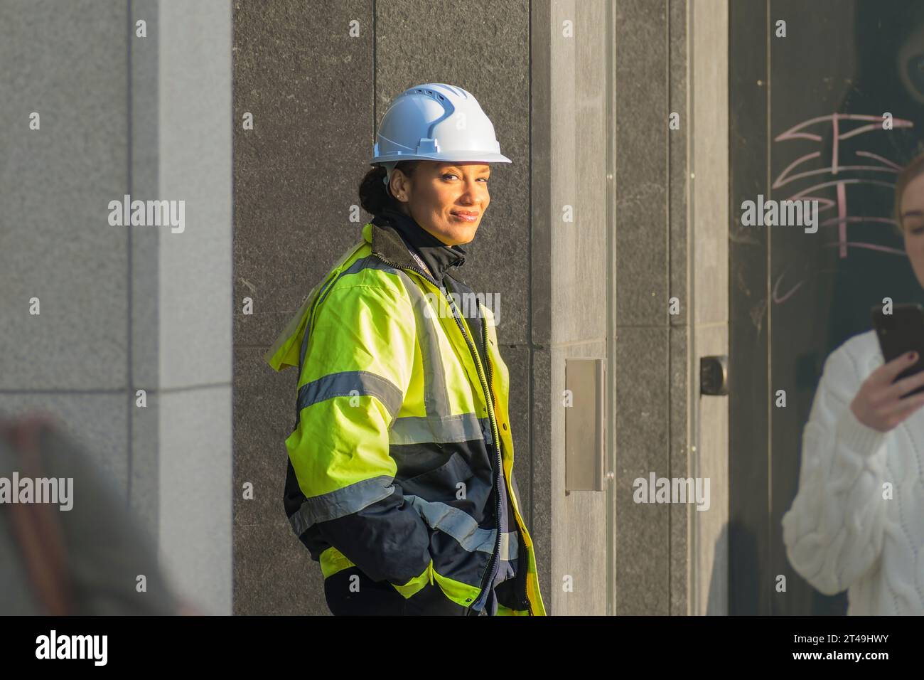 Buon costruttore. Una giovane donna con giacca Hi-Viz e casco protettivo da costruzione sulla strada di Dublino che guarda la macchina fotografica con un sorriso amichevole Foto Stock