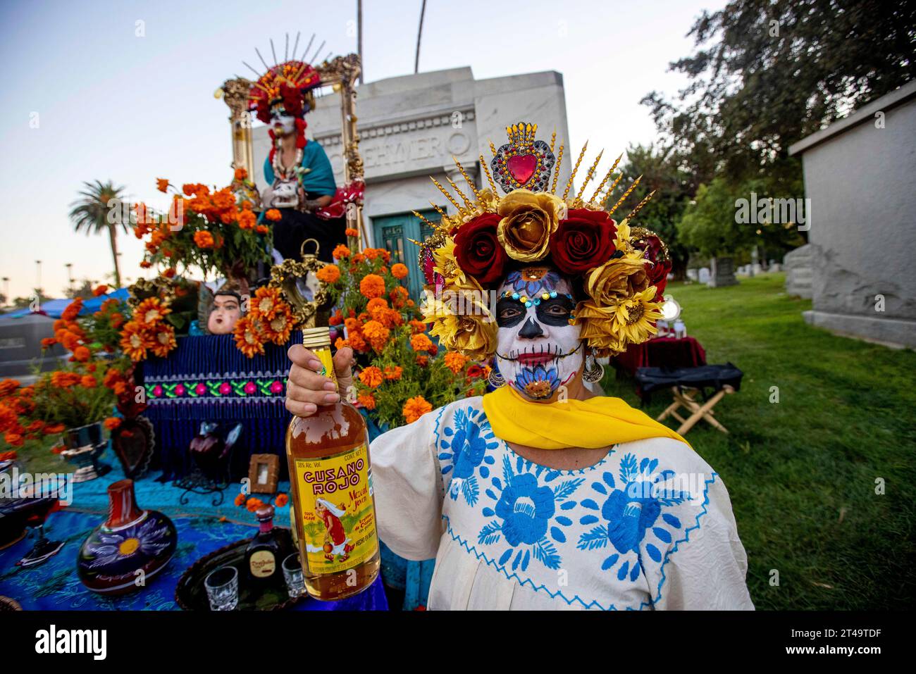 Los Angeles, California, USA. 28 ottobre 2023. Sandra Ramirez tiene una bottiglia d'epoca di mescal, che fa parte del suo altare, mentre sua figlia Xochitl Ramirez (seduta in mostra) rappresenta sua nonna e le due persone che ha perso durante la sua vita nella "Reina de mi Corazon" (Regina del mio cuore) come parte dell'evento Noche De los Muertos a Hollywood Forever nell'area di Hollywood di Los Angeles, California, il 28 ottobre 2023. (Immagine di credito: © Jill Connelly/ZUMA Press Wire) SOLO USO EDITORIALE! Non per USO commerciale! Foto Stock