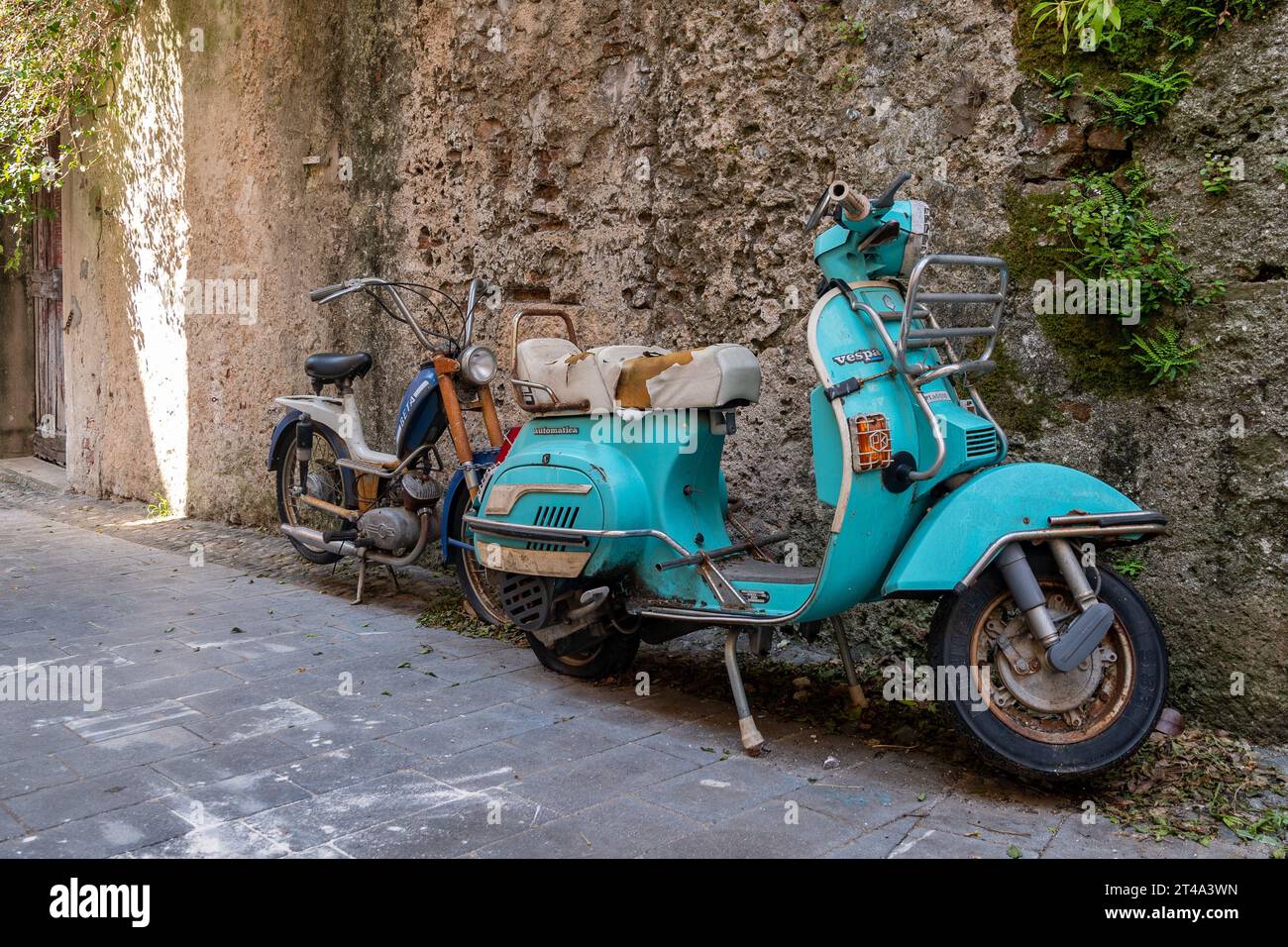 Un ciclomotore Beta arrugginito e una vecchia Vespa Piaggio parcheggiata contro un muro di pietra in uno stretto vicolo del borgo medievale, Finalborgo, finale Ligure Foto Stock