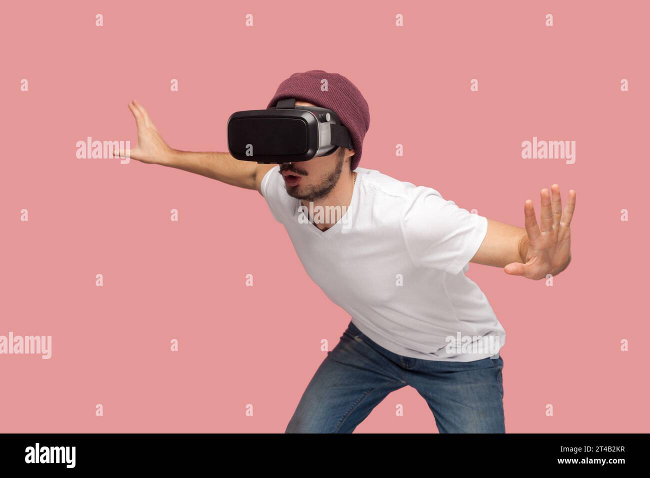 Ritratto di un uomo sbalordito e sgarbato con maglietta bianca e cappello di fagiolo, che indossa la realtà virtuale, gioca con il visore, cercando di evitare il mostro nei videogiochi. Riprese in studio per interni isolate su sfondo rosa. Foto Stock
