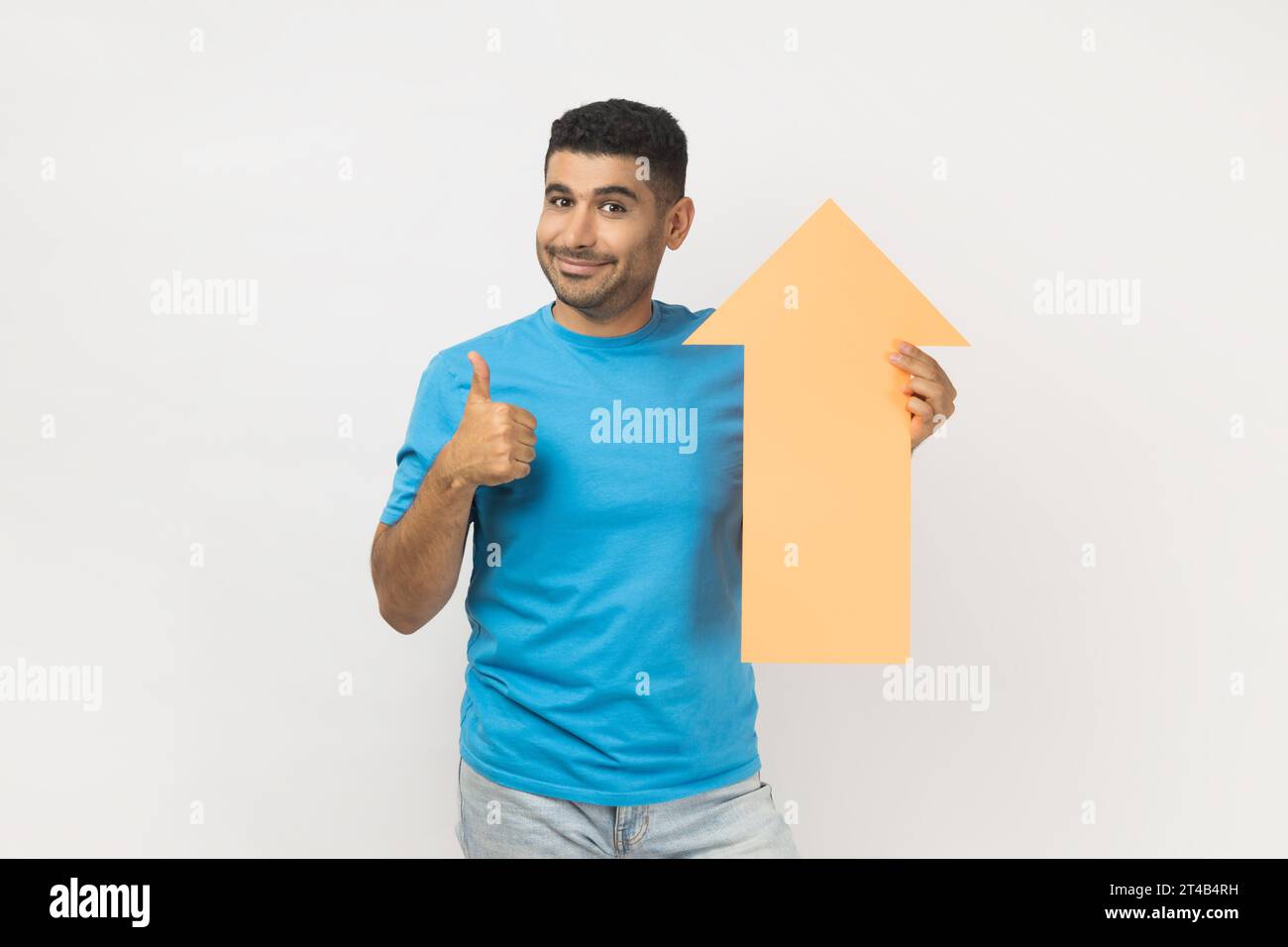 Ritratto di un uomo allegro e non rasato che indossa una T-shirt blu che tiene in piedi la freccia di cartone che indica in alto, che è felice del successo della carriera, che mostra il pollice in alto. Riprese in studio in interni isolate su sfondo grigio. Foto Stock
