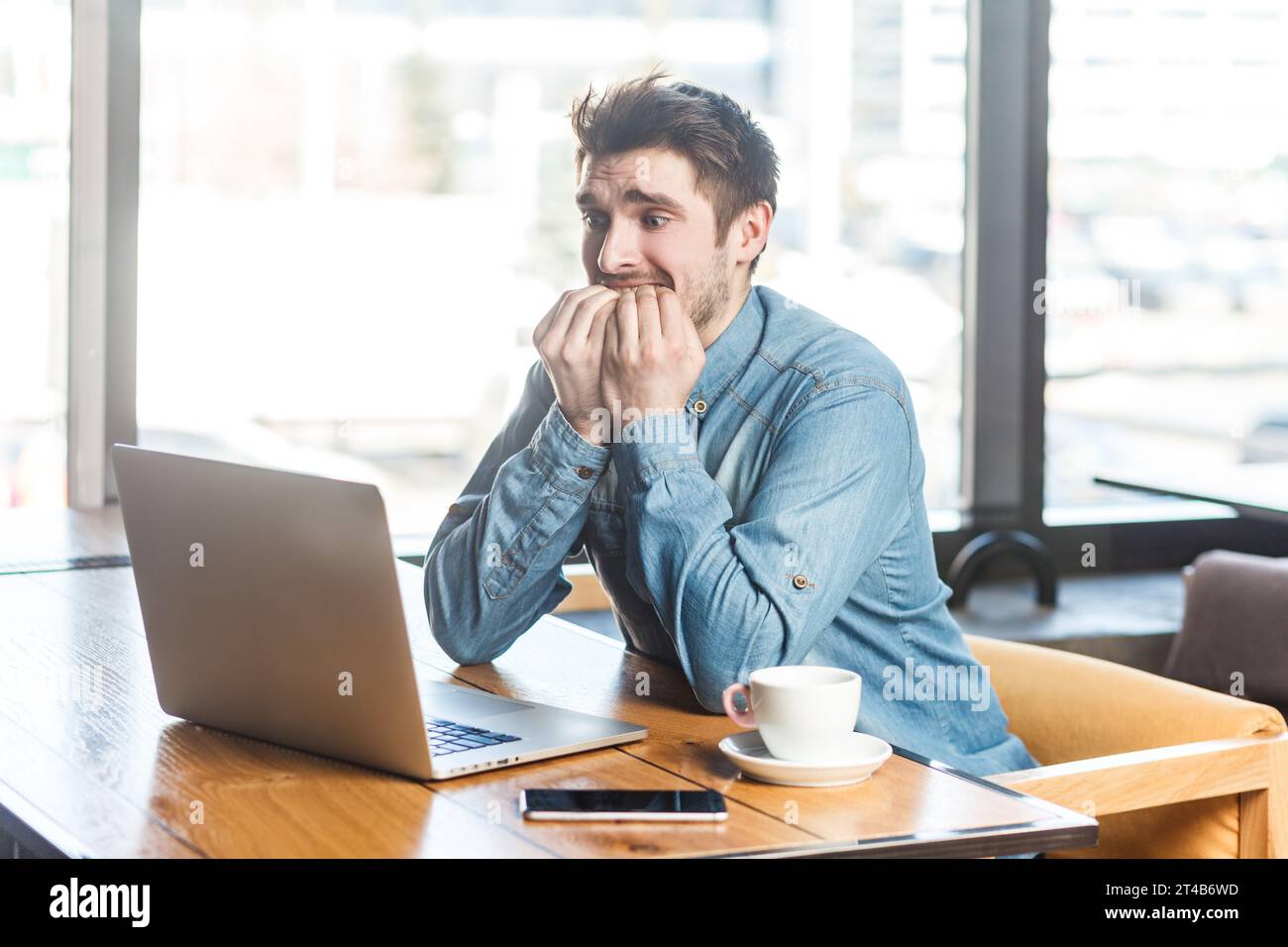 Giovane freelance nervoso preoccupato con camicia Blue jeans che lavora su un laptop, guarda lo schermo, morde le unghie delle dita, in attesa dei risultati dell'intervista. Foto al coperto vicino a una grande finestra, sfondo del caffè. Foto Stock