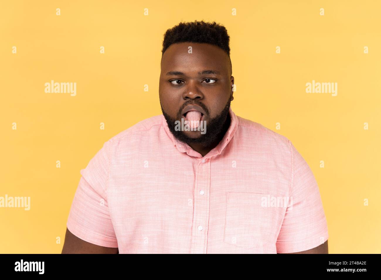 Ritratto di un uomo disobbediente, pazzo e divertente che indossa una camicia rosa che mostra la lingua e gli incrocia gli occhi scontento, comportandosi in modo indebito e cattivo. Riprese in studio in interni isolate su sfondo giallo. Foto Stock
