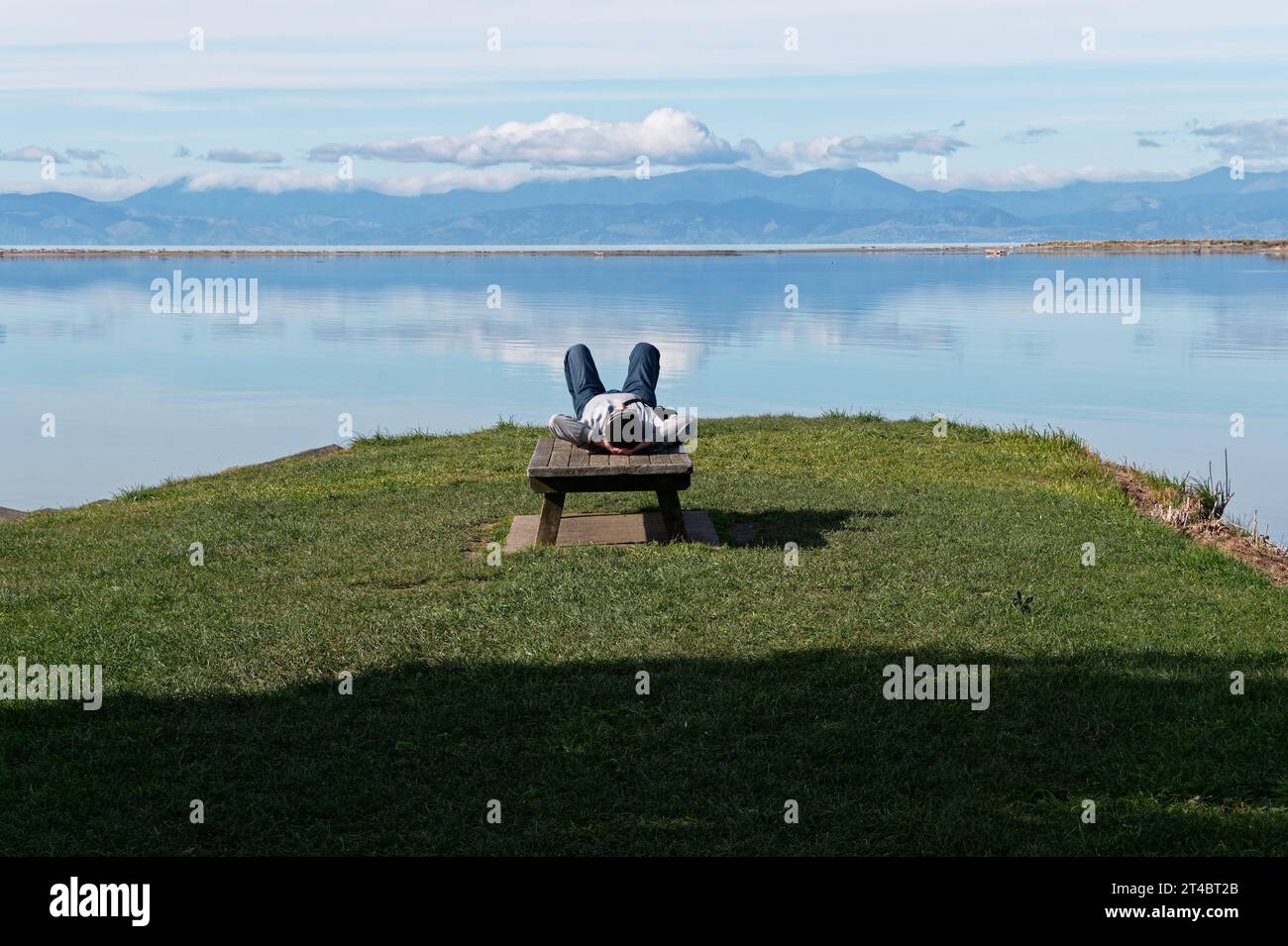 Una persona giace sulla schiena al molo storico di Motueka. La Motueka Sandspit può essere vista con Nelson sullo sfondo. Foto Stock