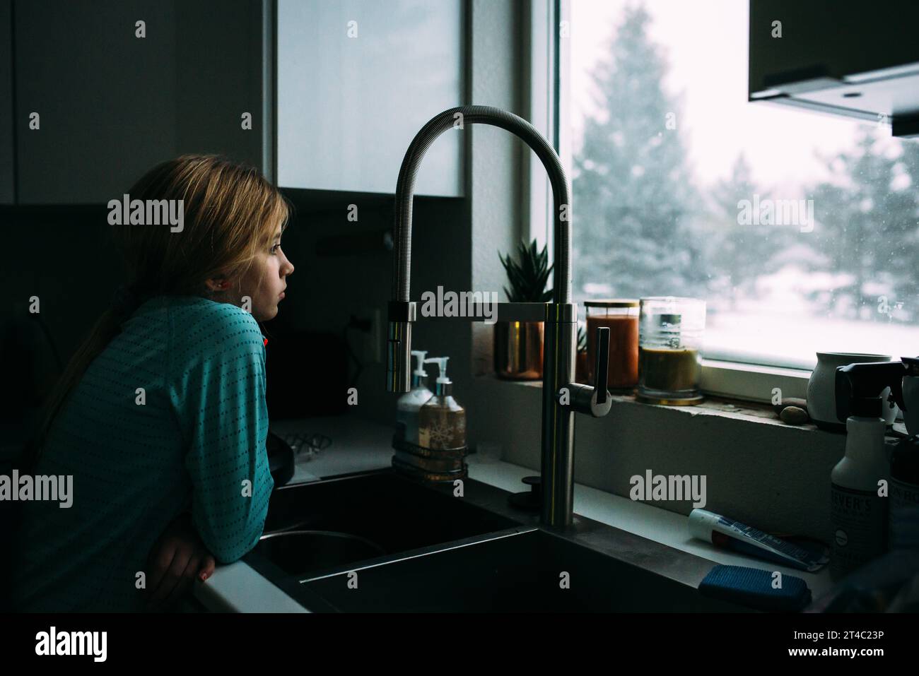 La ragazza preadolescente si appoggia sul lavandino della cucina e guarda fuori dalla finestra di sno Foto Stock