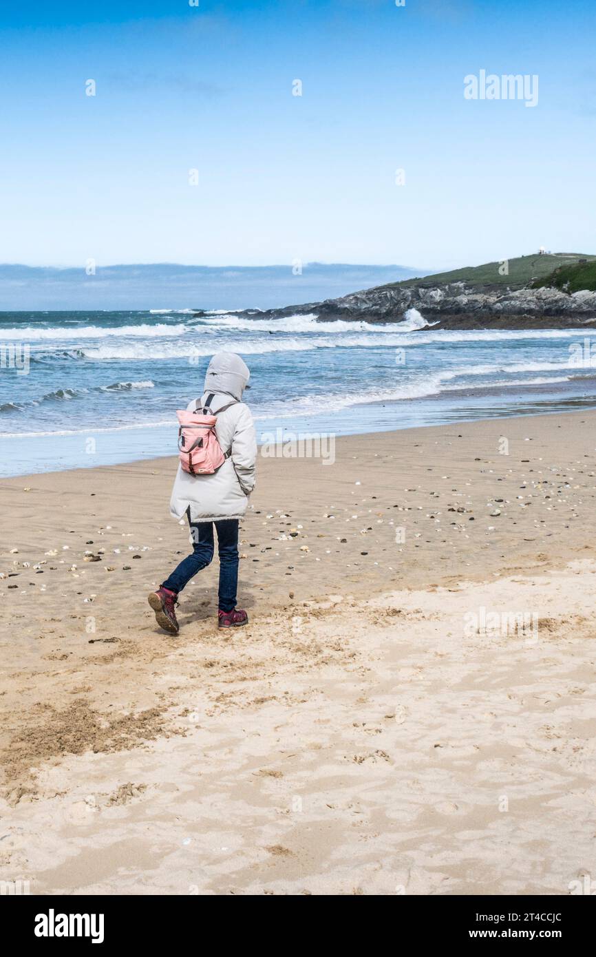 Una donna che indossa un cappotto caldo con un cappuccio che cammina lungo Fistral Beach in fresco sole primaverile a Newquay in Cornovaglia nel Regno Unito. Foto Stock