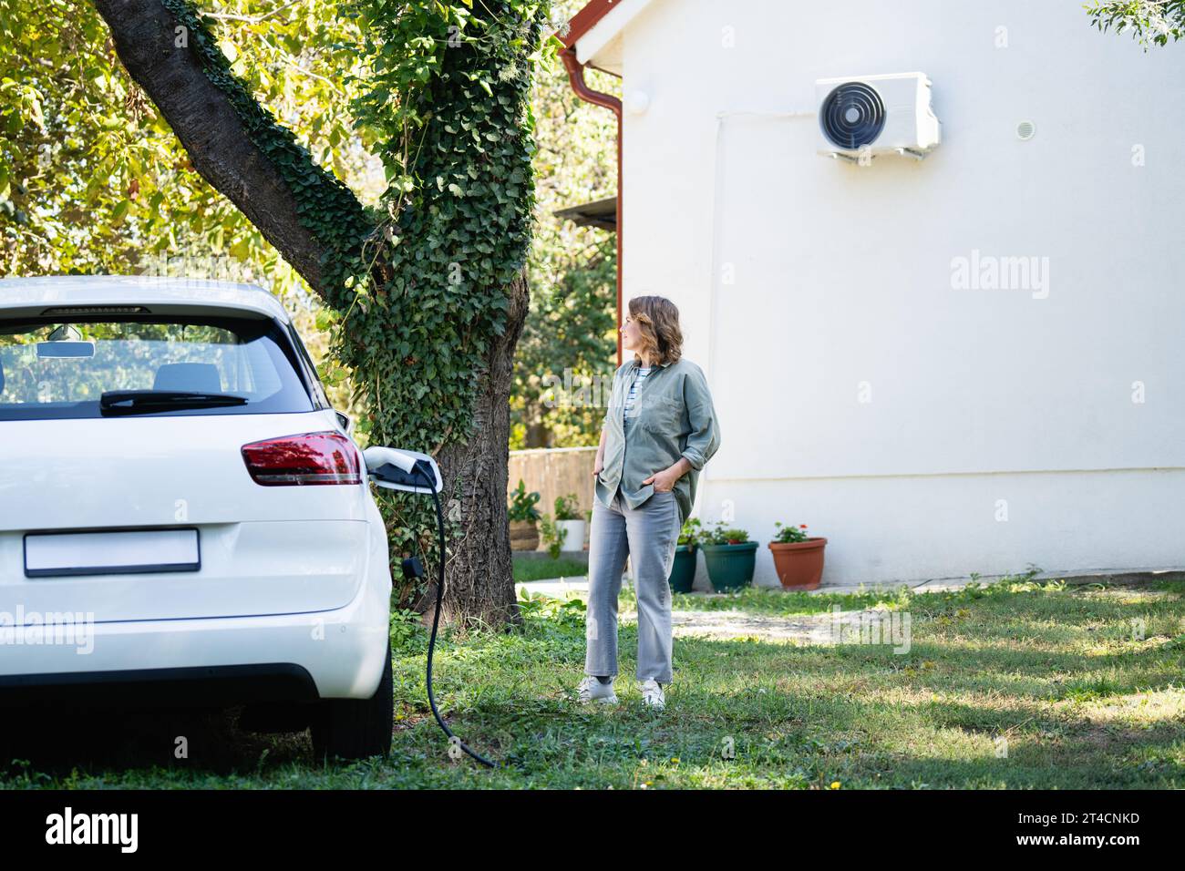 Donna vicino a un'auto elettrica in carica nel cortile di una casa di campagna. Foto Stock