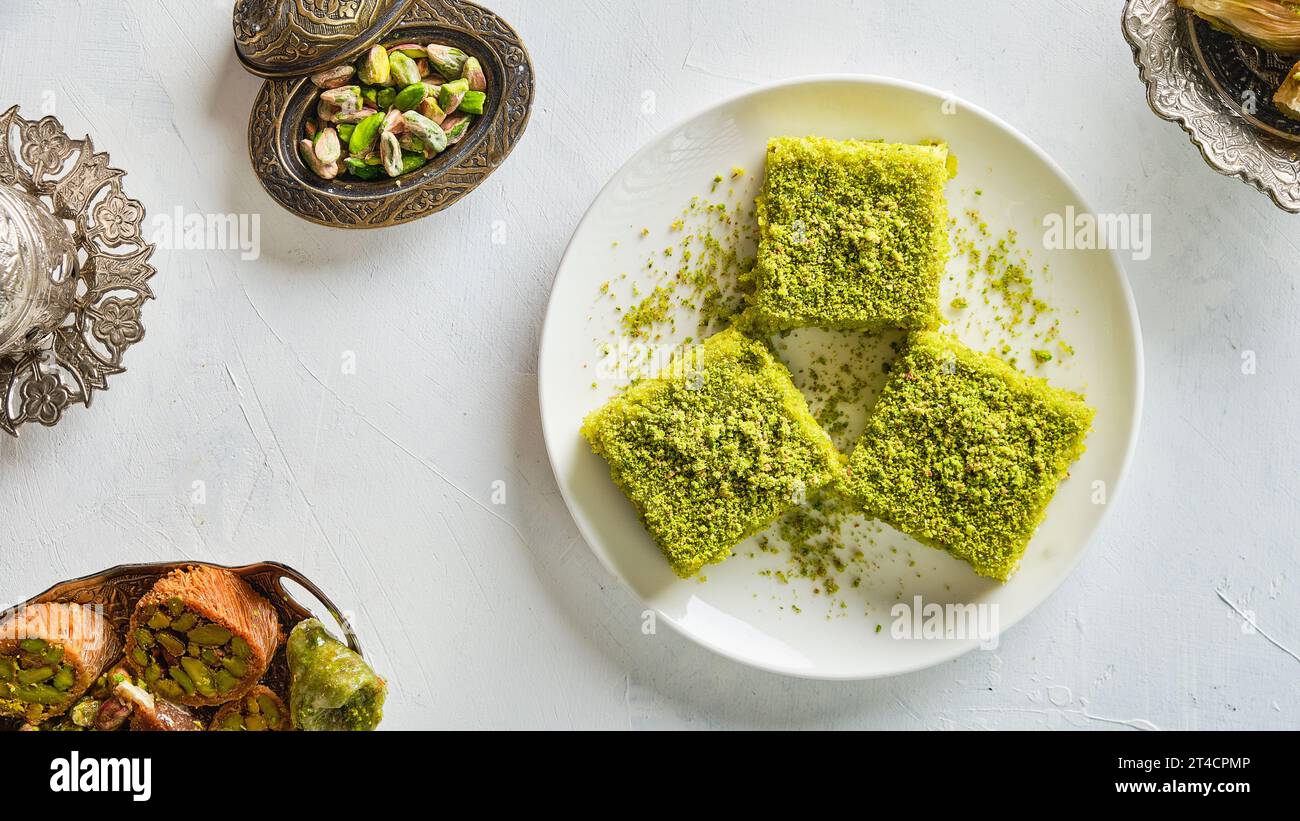 Pezzi di dolci orientali con pistacchi in un piatto bianco su un terreno bianco circondato da utensili in argento e rame Foto Stock