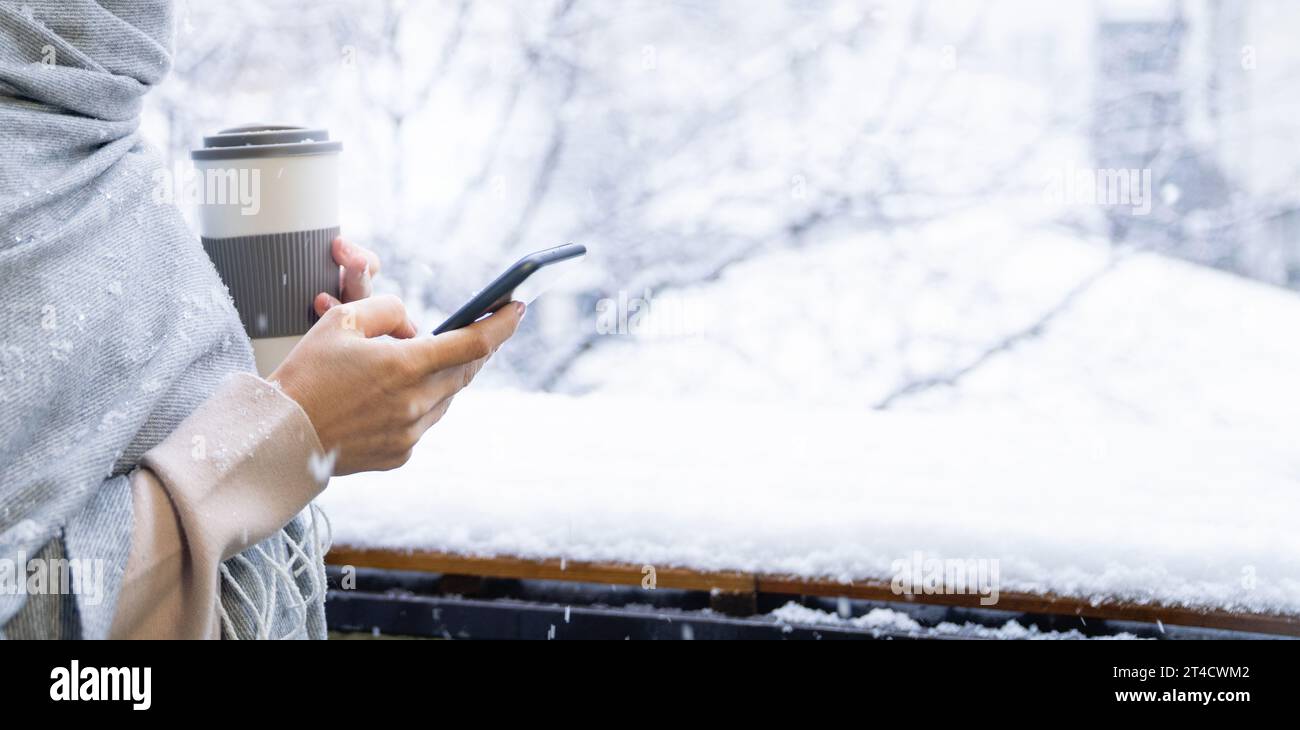 Una donna con un caldo cappello e una coperta tiene lo smartphone tra le mani. Paesaggio invernale con nevicate sullo sfondo. Foto Stock
