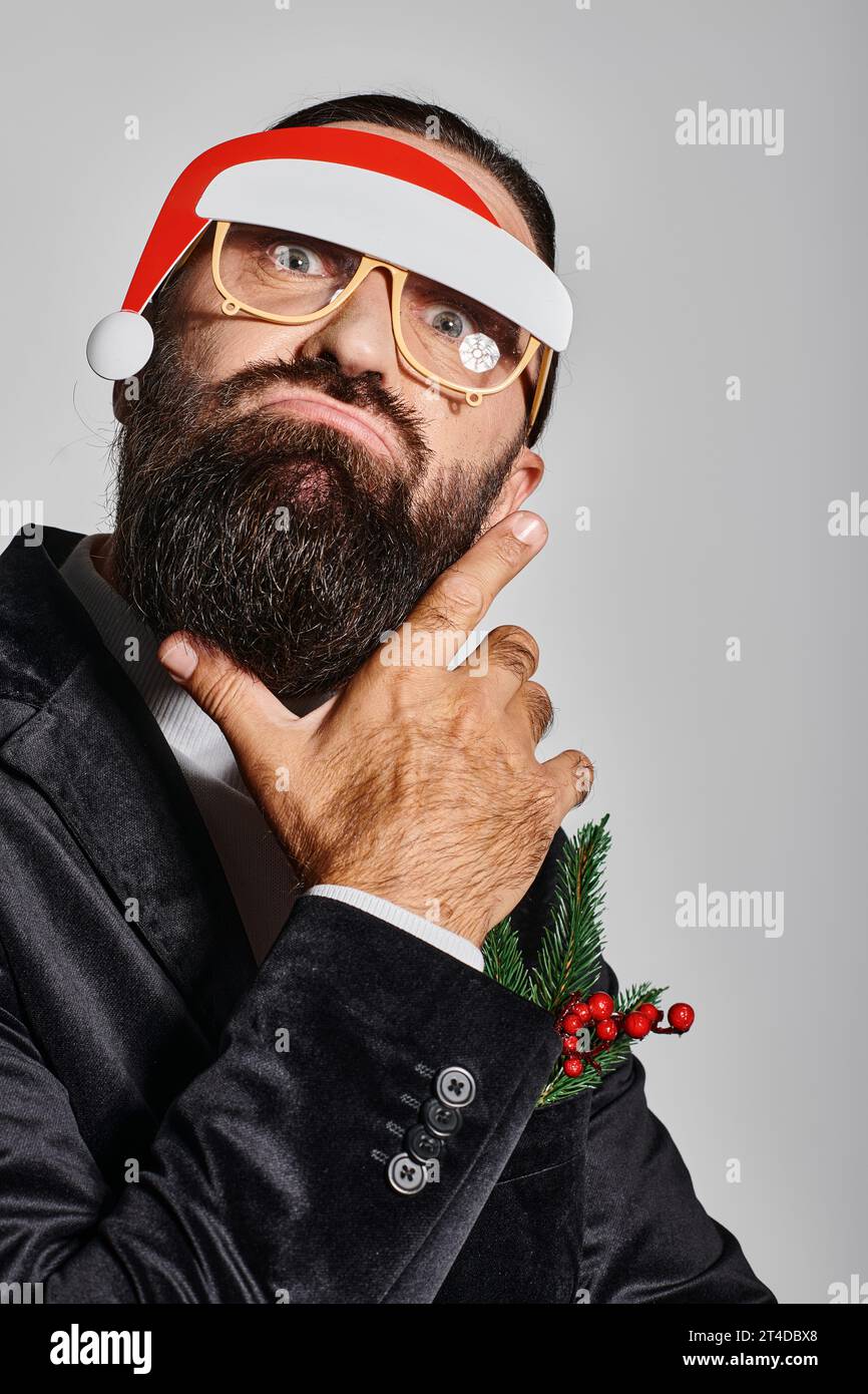 uomo barbuto con occhiali festivi con cappello di babbo natale che posa in abito e che poggia le labbra su sfondo grigio Foto Stock