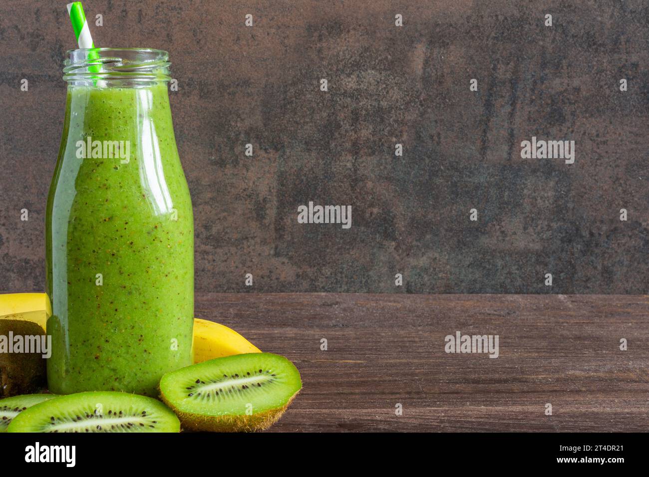 Frullato verde a base di kiwi e banana in bottiglia di vetro con frutta fresca su fondo rustico di legno. Dieta sana bevanda Foto Stock