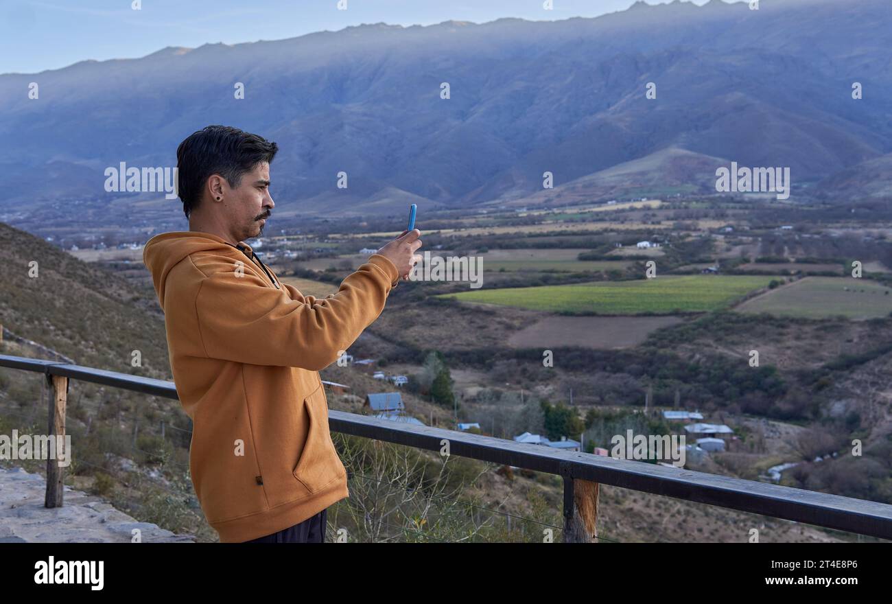 uomo latino-americano marrone vestito con una felpa con cappuccio color senape che scatta una foto con il suo cellulare di un incredibile paesaggio di valle e montagna Foto Stock