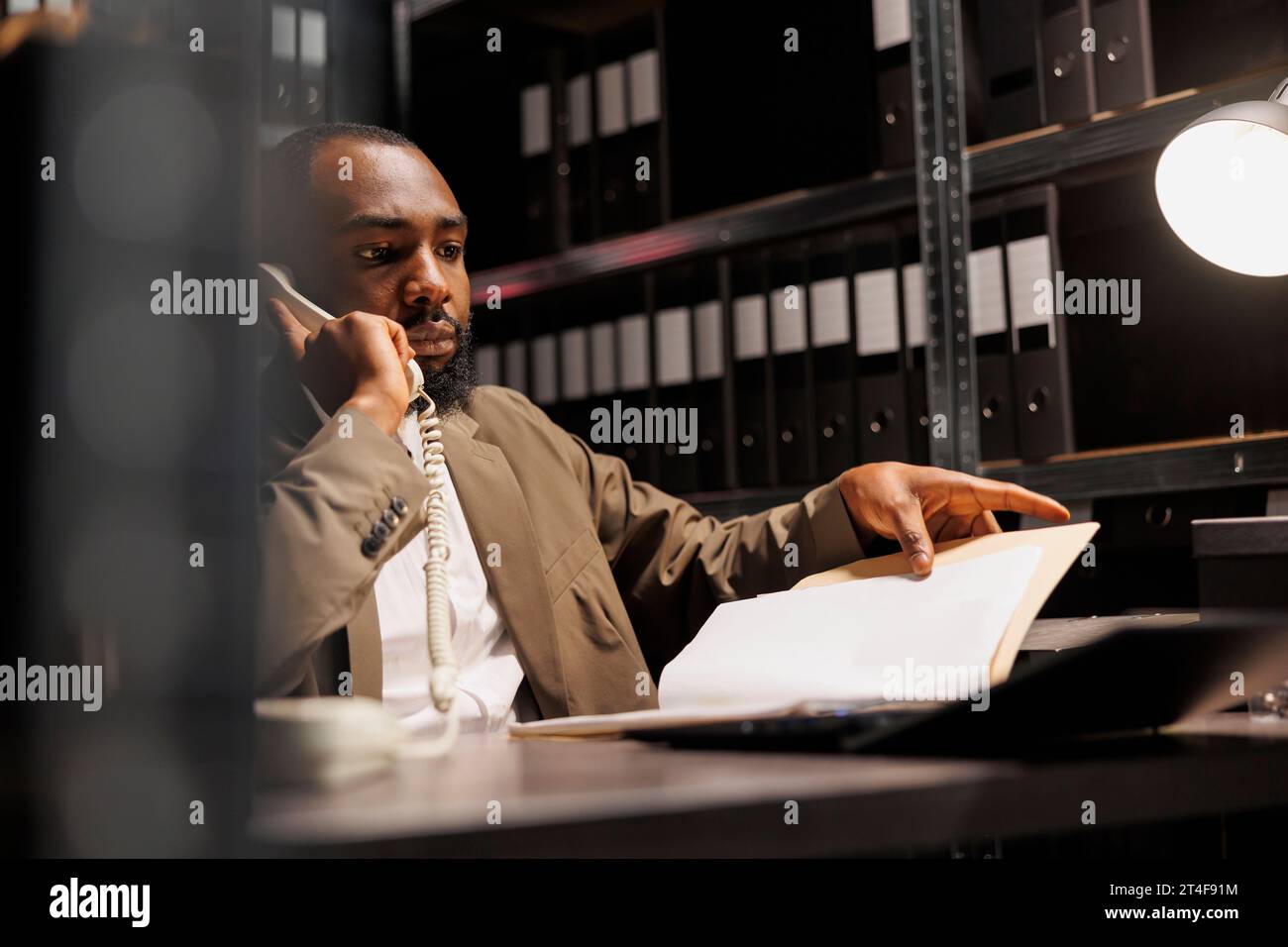 Investigatore della polizia afroamericana che risponde alle telefonate su rete fissa in ufficio di notte. Investigatore preoccupato che discute di informazioni sui casi di crimine mentre analizza il file del rapporto forense Foto Stock