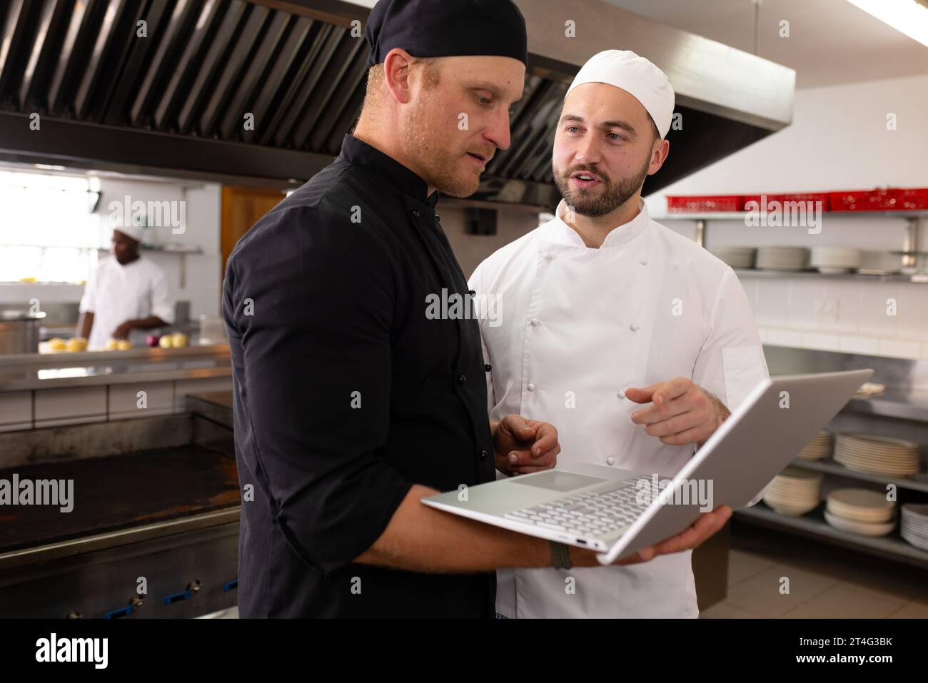 Insegnante di cuoco maschio caucasico che usa un computer portatile mentre discute con uno studente in cucina commerciale Foto Stock