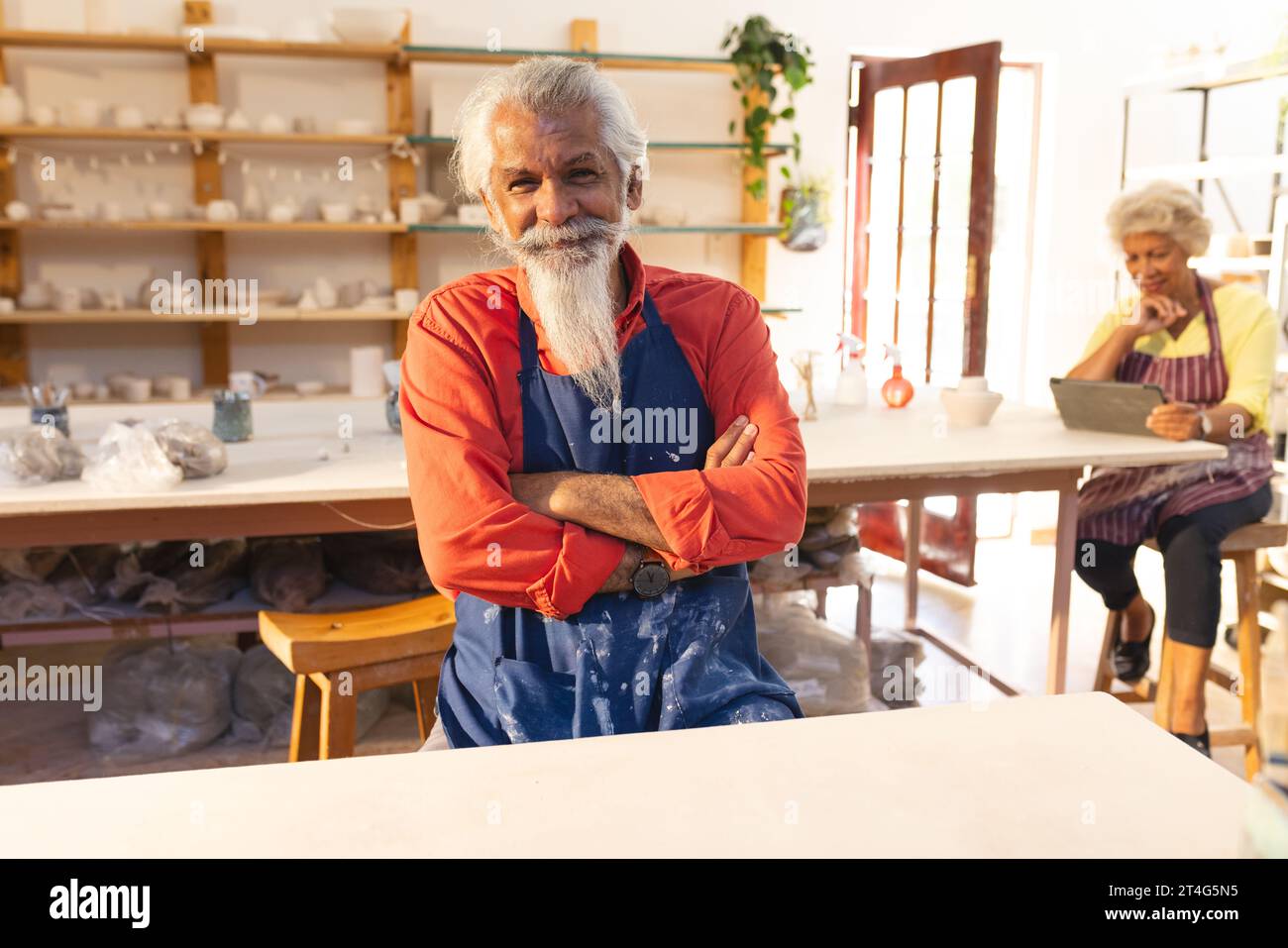 vasaio esperto birazziale con barba lunga seduto e sorridente nello studio di ceramica Foto Stock