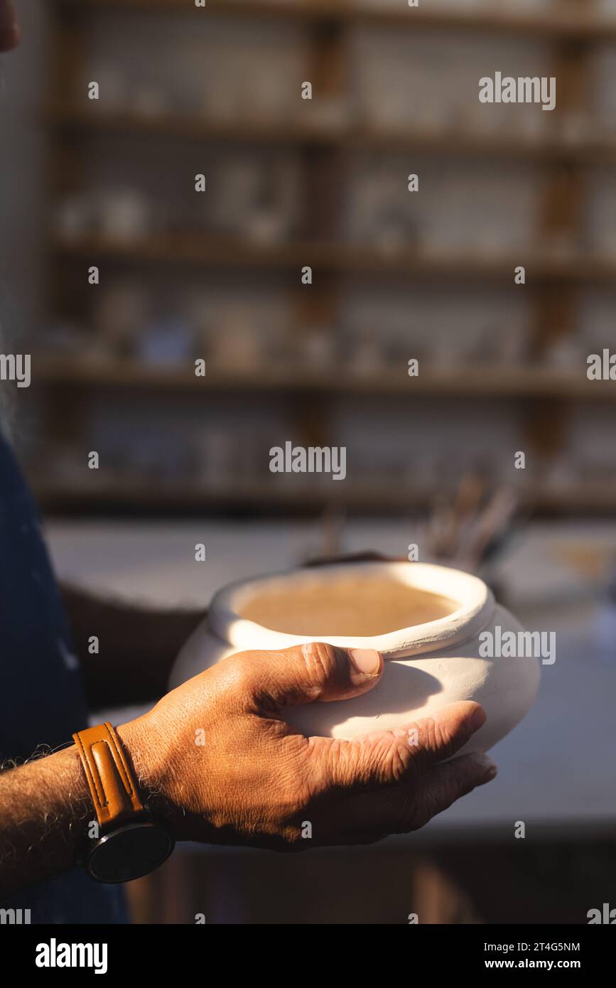 Primo piano delle mani di un vasaio birazziale anziano che tiene un vaso di argilla nello studio di ceramica Foto Stock