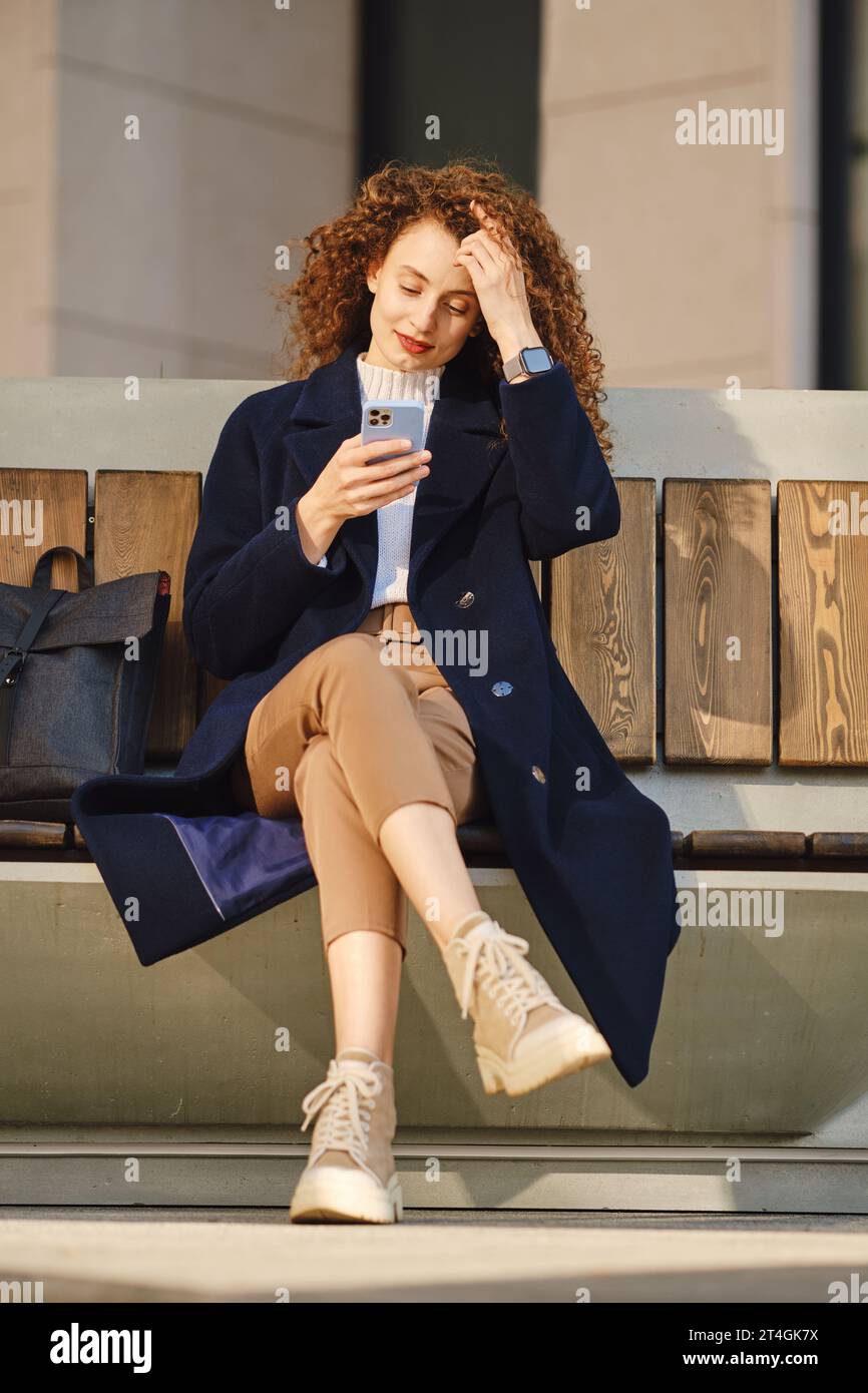 Donna in cappotto siede sulla panchina in strada con lo smartphone nelle giornate di sole Foto Stock