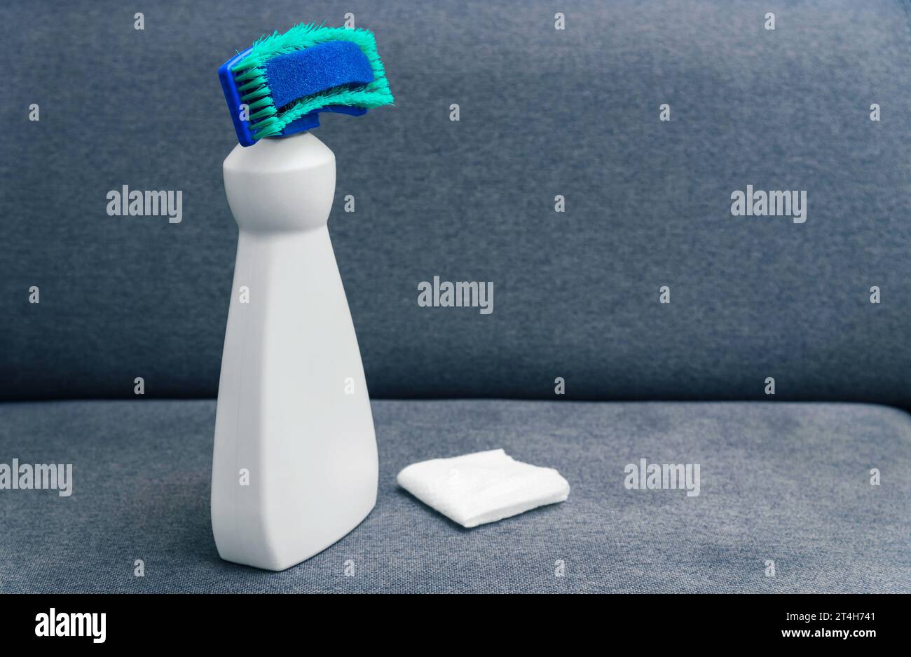 Prodotto per la pulizia con spugna. Smacchiatore per tessuti per mobili. Bottiglia bianca in plastica con detergente e tovagliolo su un divano grigio. Foto Stock