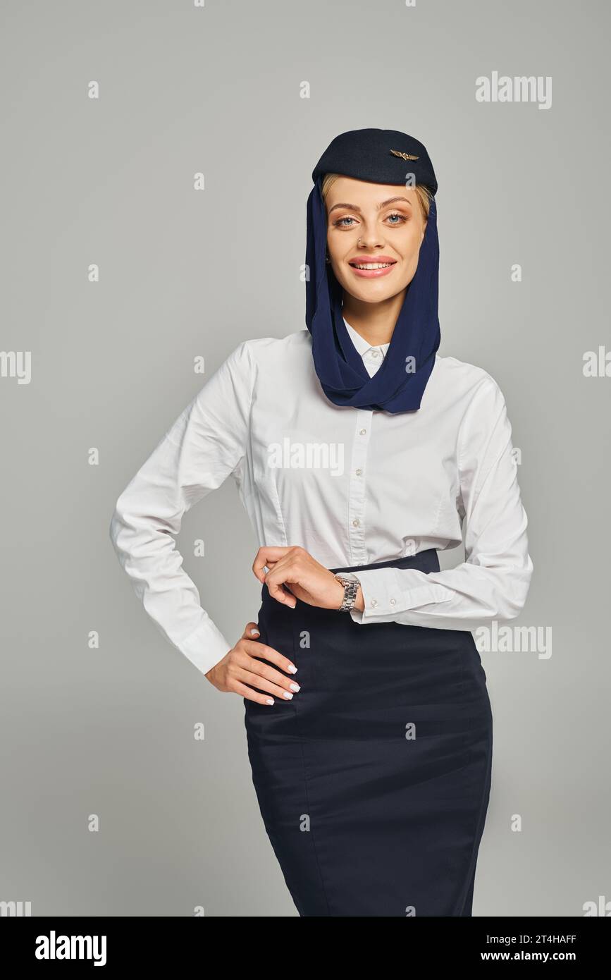 elegante e gioiosa hostess in uniforme delle compagnie aeree arabe in piedi con la mano sul fianco del grigio Foto Stock