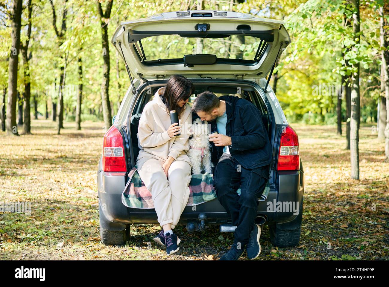 Adora la giovane famiglia accarezza e baciare il suo carino cane mentre si siede nel bagagliaio dell'auto e si rilassa nella foresta. Concetto di famiglia, amore, vacanza e viaggio Foto Stock