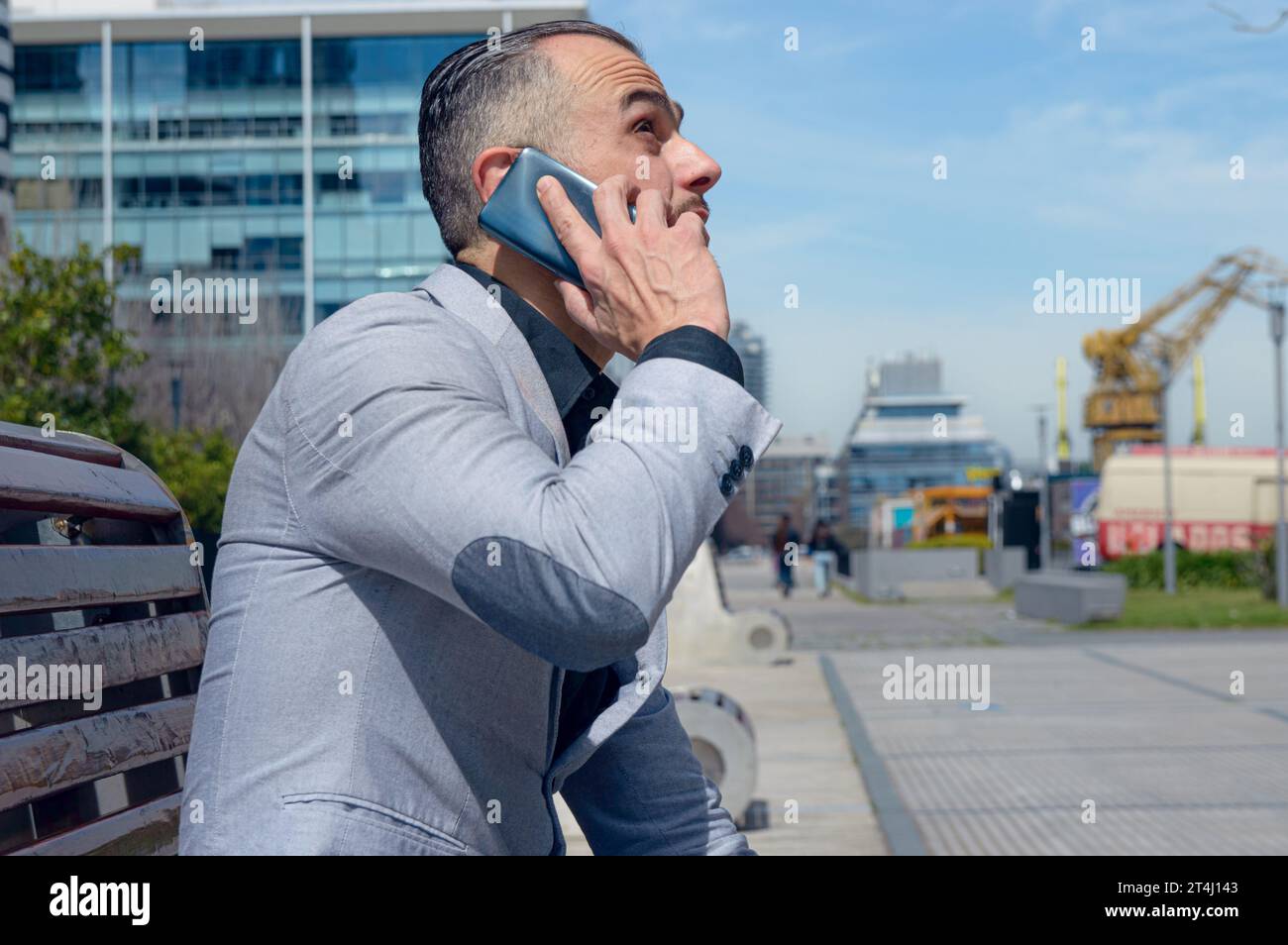 uomo d'affari caucasico adulto, indossa un blazer grigio, capelli corti, è seduto all'aperto a parlare al telefono guardando il cielo e attento e attento all'ascolto Foto Stock