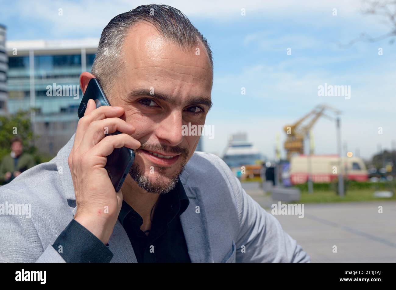 Ritratto di un uomo d'affari caucasico adulto felice con giacca grigia e capelli corti, sorridente, seduto a parlare al telefono all'aperto, guardando la macchina fotografica, sbirciare Foto Stock