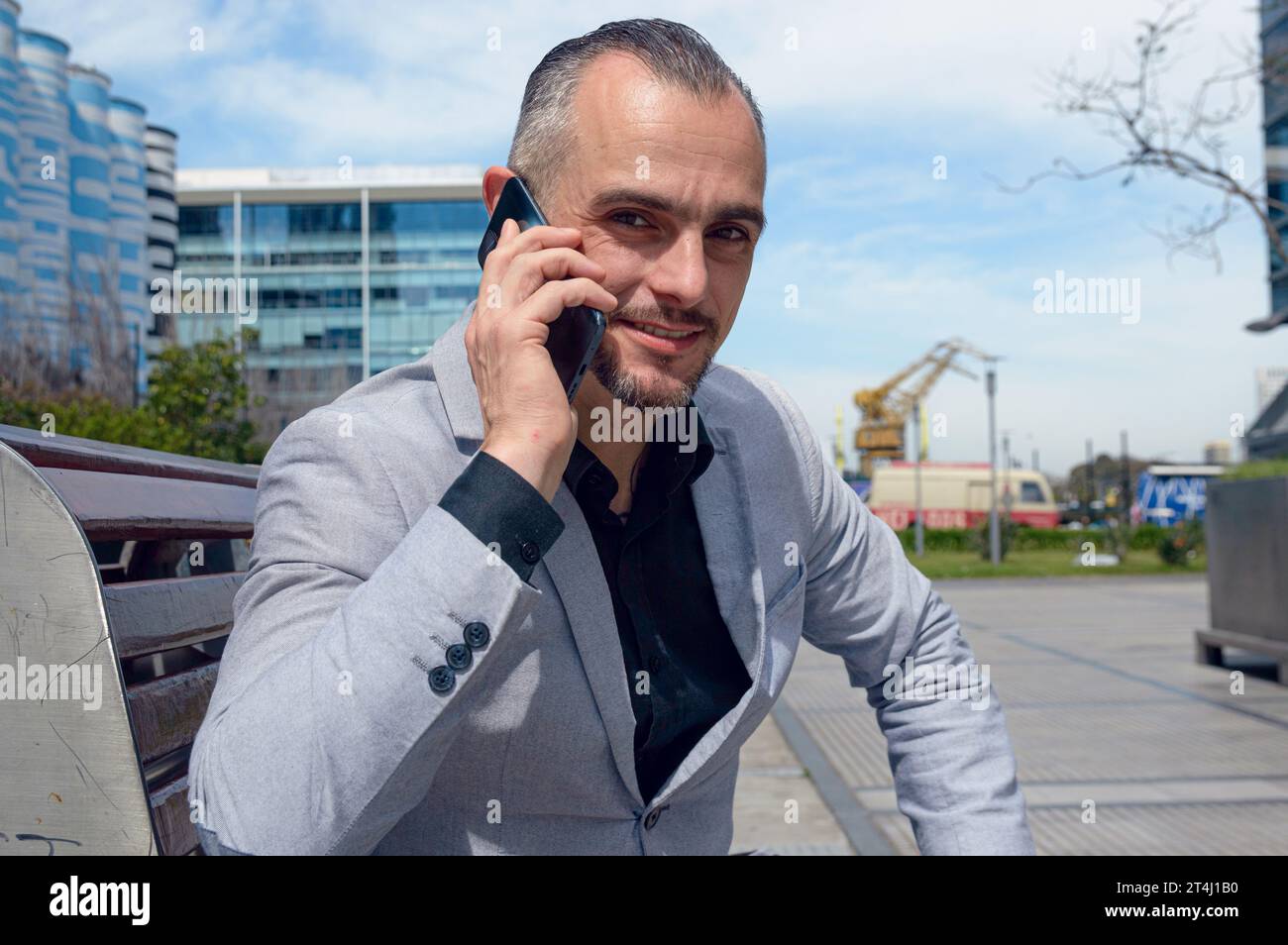 Ritratto di un uomo d'affari caucasico adulto felice con giacca grigia e capelli corti, sorridente, seduto a parlare al telefono all'aperto, guardando la macchina fotografica, sbirciare Foto Stock