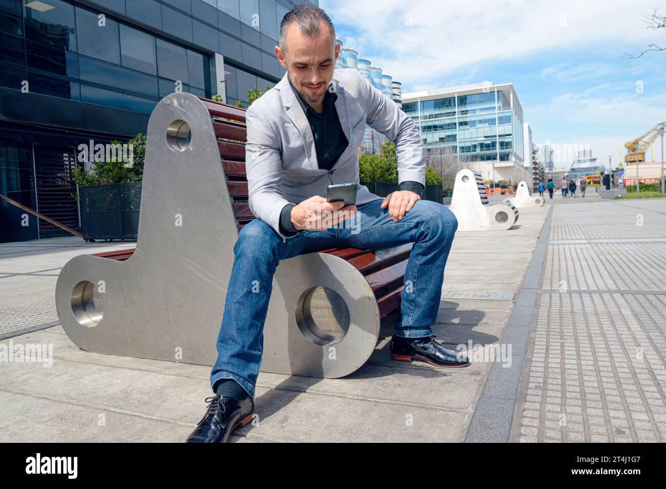 Uomo adulto caucasico di etnia francese e uomo d'affari, seduto a mezzogiorno all'aperto, felice di guardare il telefono controllare i social media e le notifiche Foto Stock
