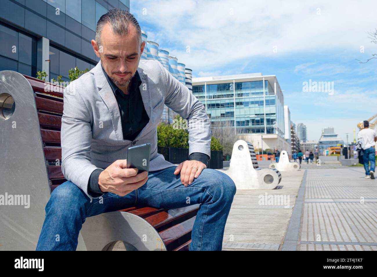 Uomo adulto caucasico di etnia francese e uomo d'affari, seduto a mezzogiorno all'aperto, felice di guardare il telefono controllare i social media e le notifiche Foto Stock