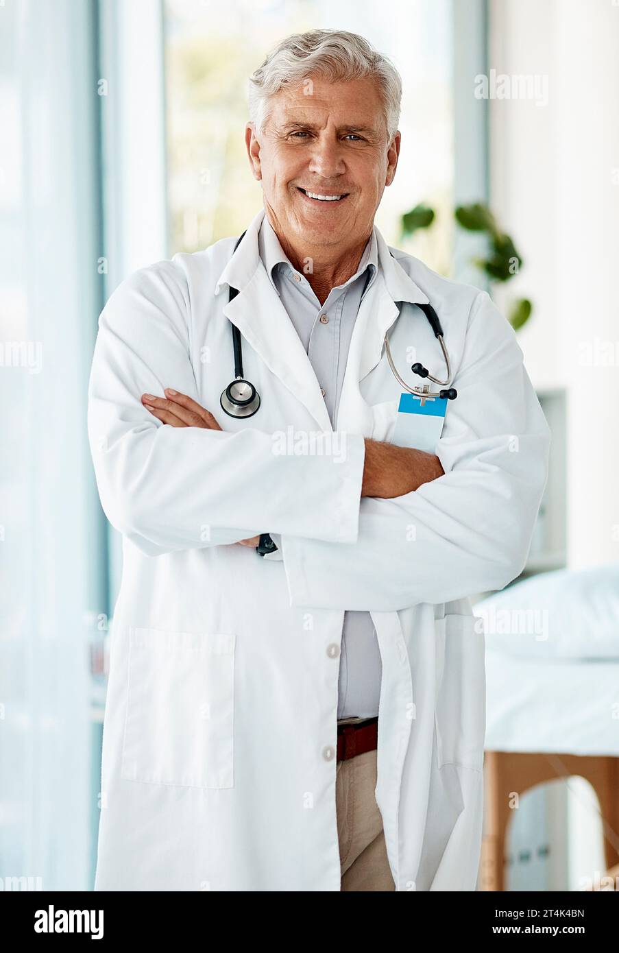 Esperto medico maschio maturo sorridente e in piedi con le braccia incrociate in un ospedale. Un uomo anziano ottimista che indossa un camice da laboratorio Foto Stock
