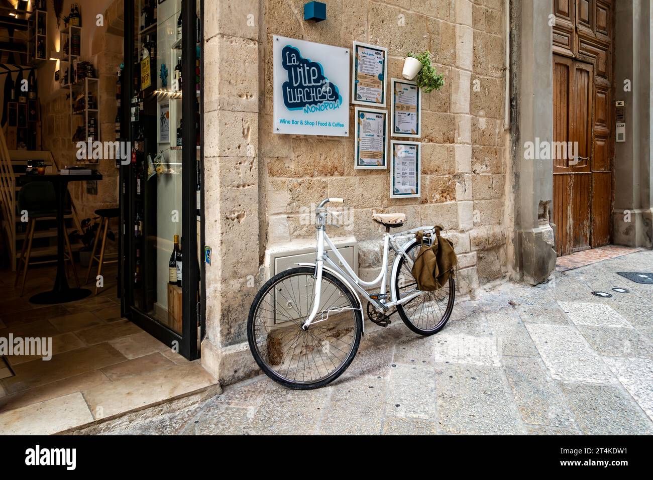 Affascinante ingresso al wine bar a Monopoli, in Italia, con una bicicletta bianca d'epoca e menu esposti su una rustica parete di pietra lungo una stretta strada acciottolata. Foto Stock
