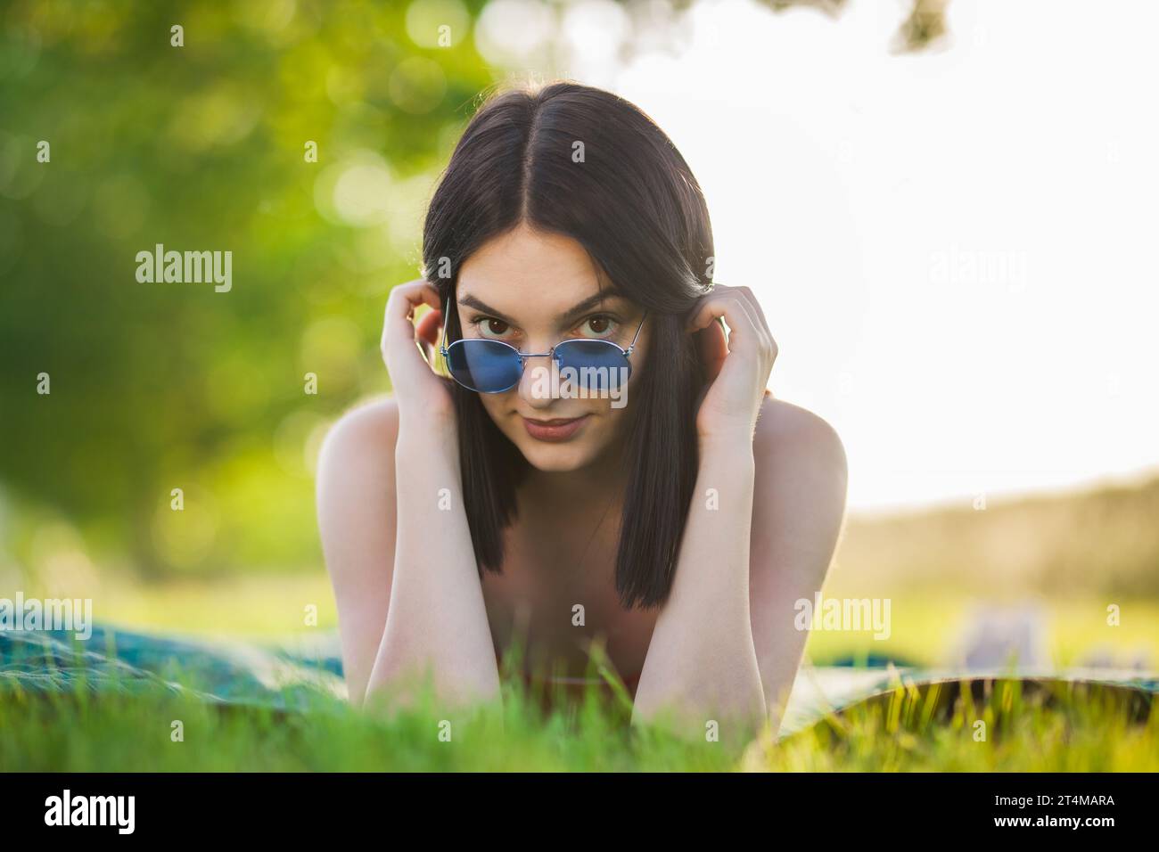 Bellissima ragazza dai capelli scuri con occhiali da sole sdraiata su un prato nel parco. Guardando la fotocamera. Foto Stock