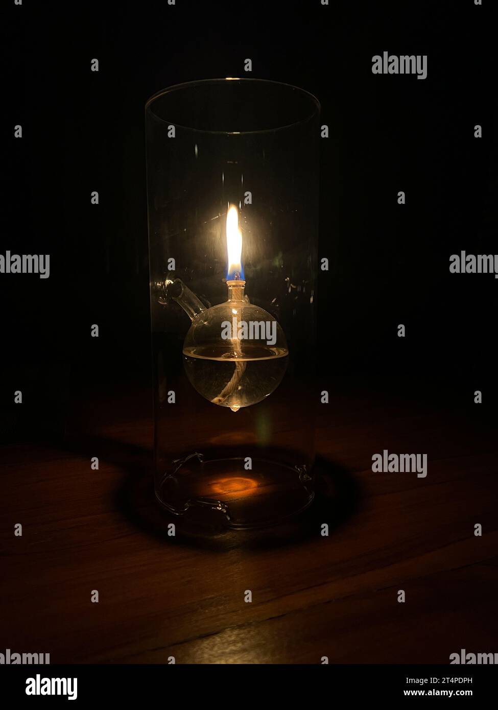 Lampada a olio in vetro trasparente illuminata posizionata su un tavolo al buio creando un bagliore con sfondo scuro. Foto Stock