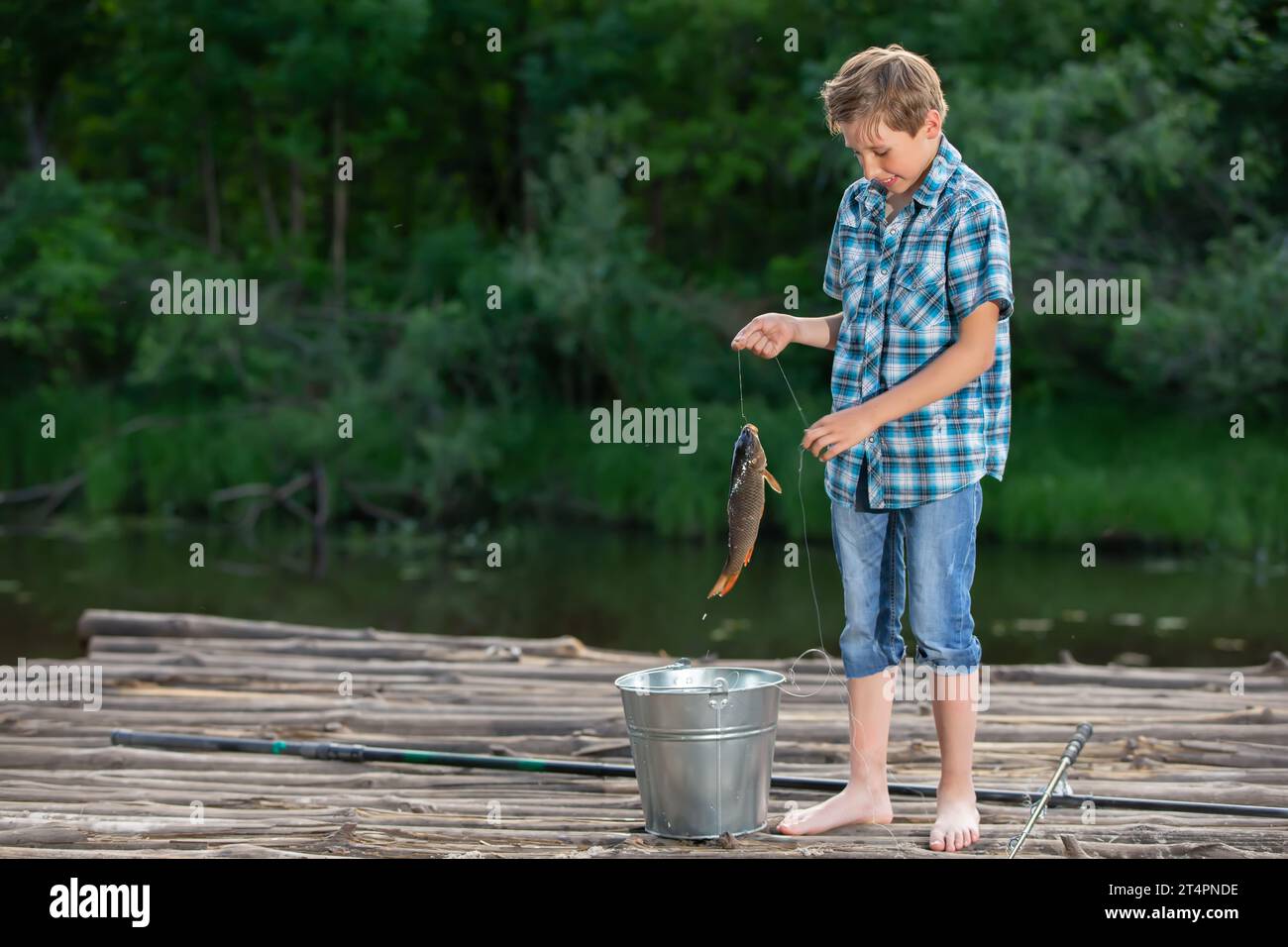 Un ragazzo di pescatori si trova su un ponte di legno e guarda la carpa catturata. Foto Stock