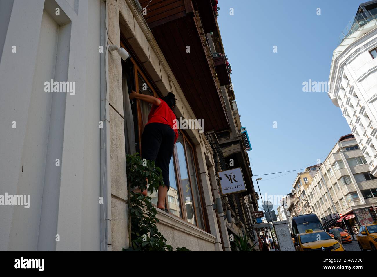 La donna di colore rosso sgancia senza sforzo fuori dalla finestra della hall dell'hotel, abbracciando l'audace compito di pulirla in mezzo allo sfondo urbano. Foto Stock