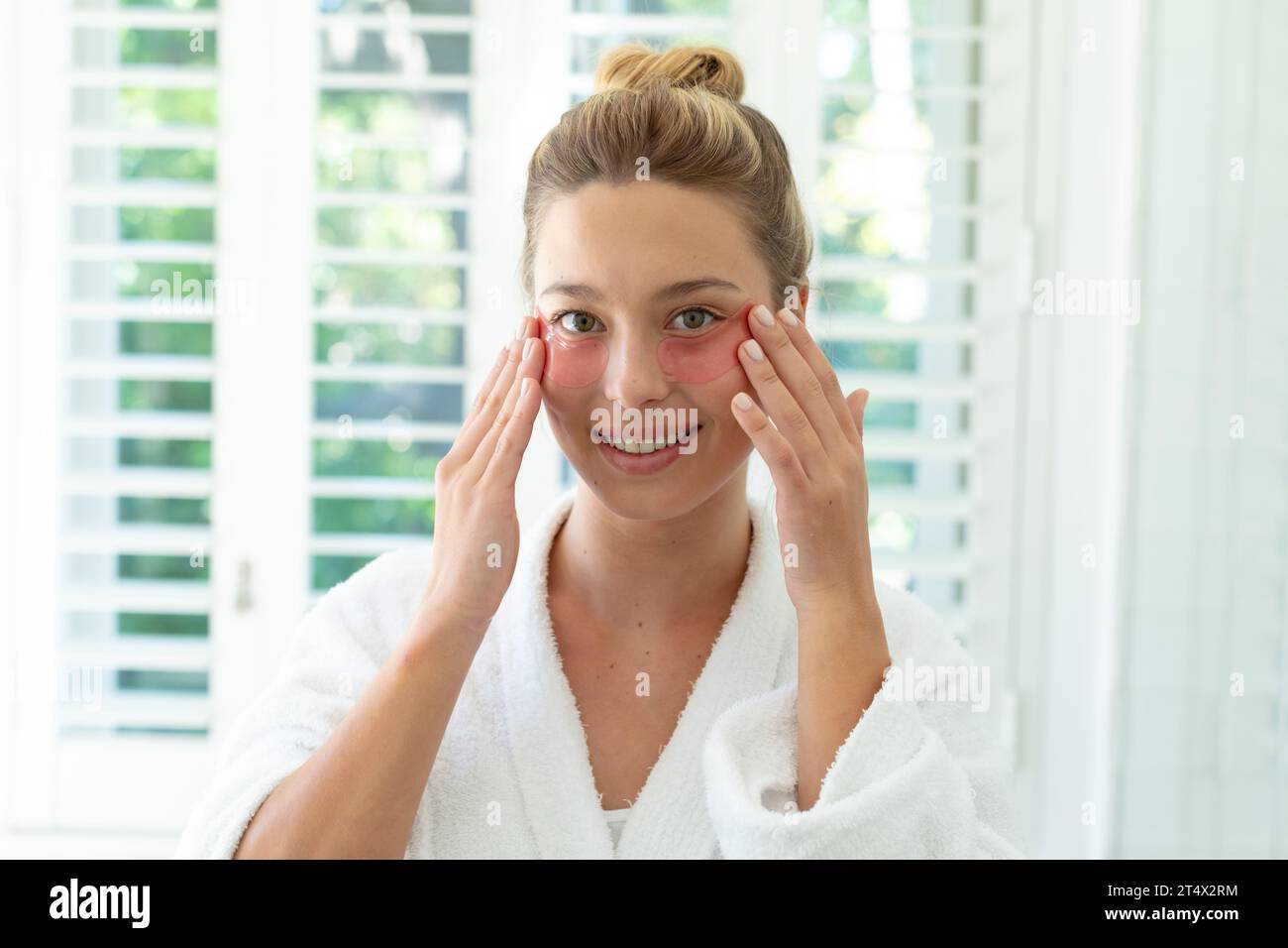 Donna caucasica felice in accappatoio che mette maschera gel sotto gli occhi nel bagno soleggiato Foto Stock