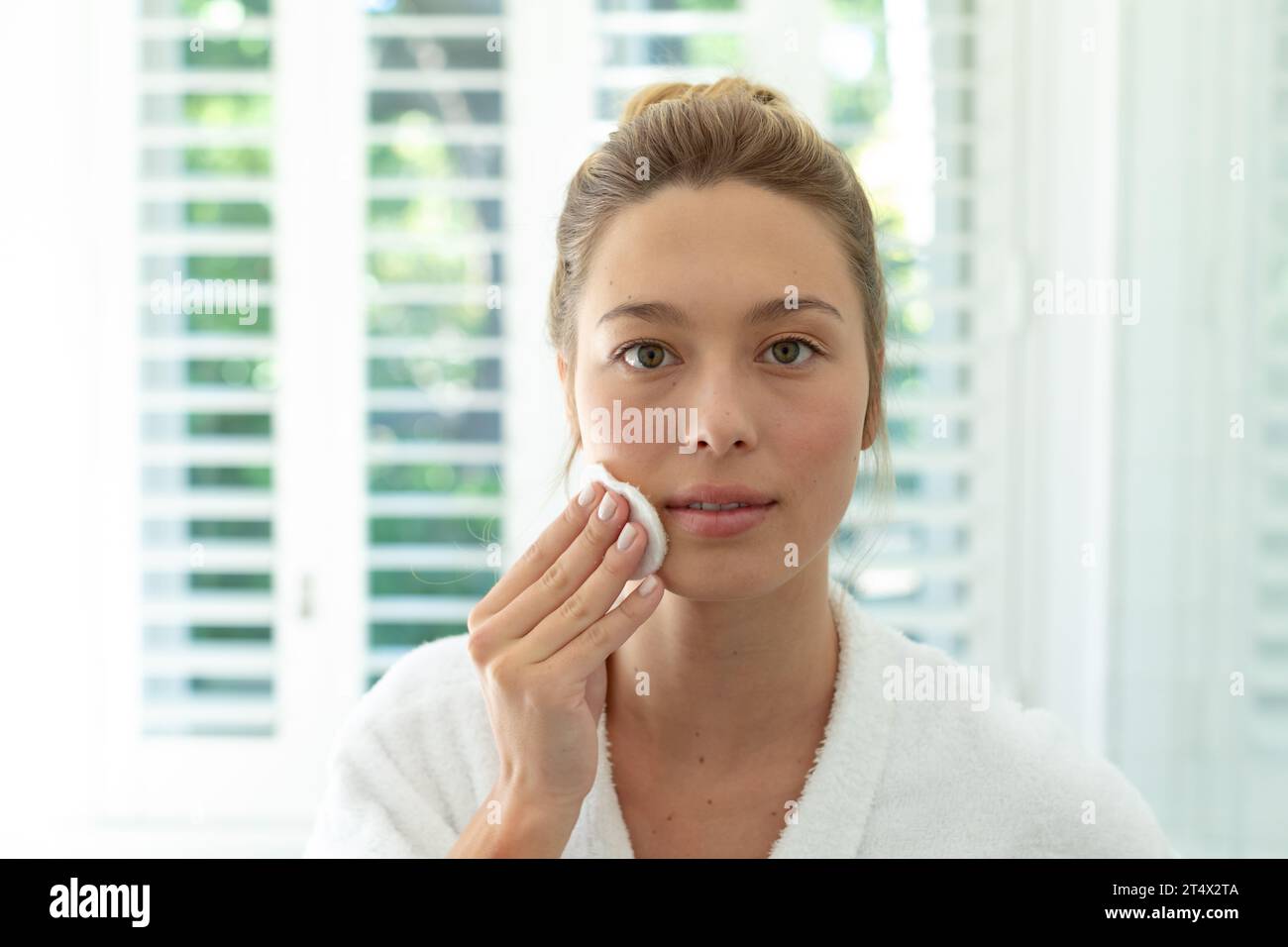 Donna caucasica felice in accappatoio che pulisce il viso con batuffolo di cotone nel bagno soleggiato Foto Stock