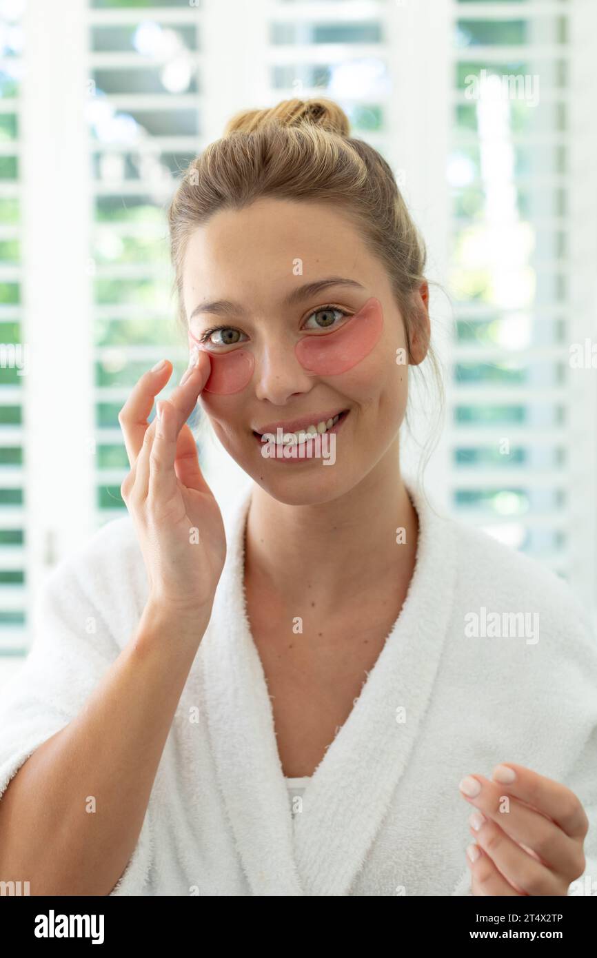 Donna caucasica felice in accappatoio che mette maschera gel sotto gli occhi nel bagno soleggiato Foto Stock