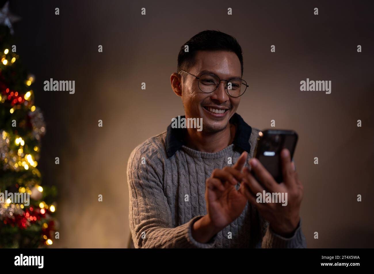 Un allegro uomo asiatico con un accogliente maglione sta chiacchierando con gli amici o inviando messaggi di Natale alla sua famiglia mentre si rilassa nel suo soggiorno su un Foto Stock