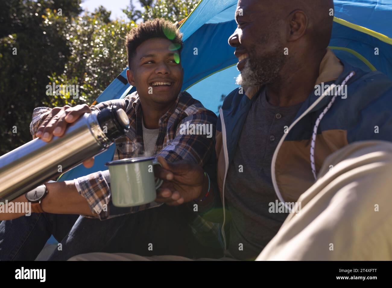 Buon padre afroamericano e figlio adulto seduti fuori dalla tenda mentre bevevano un caffè al sole Foto Stock