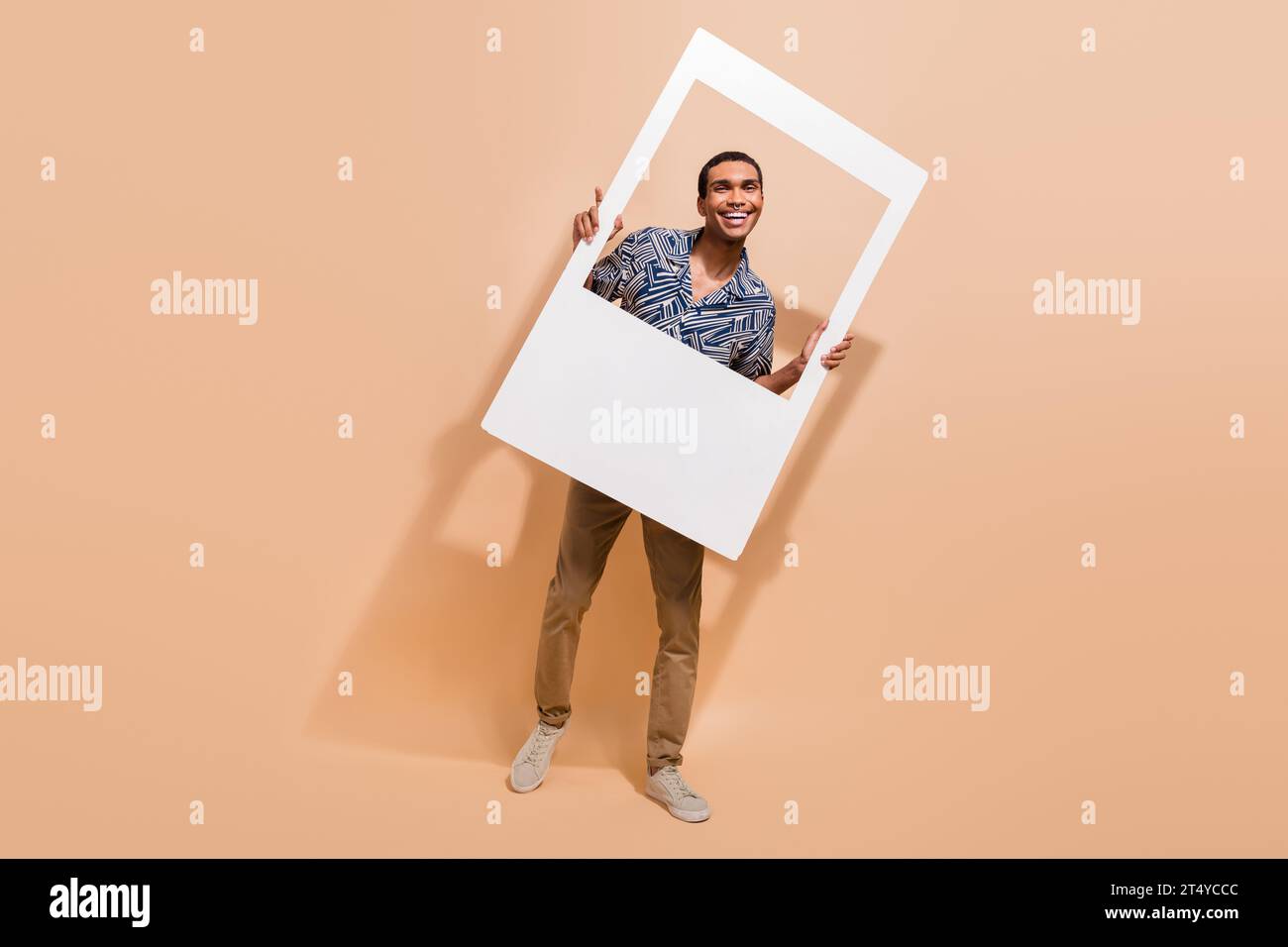 Foto a grandezza naturale di un uomo di buon umore che indossa pantaloni con stampa camicia con cornice bianca che rende l'immagine istantanea isolata su sfondo di colore beige Foto Stock