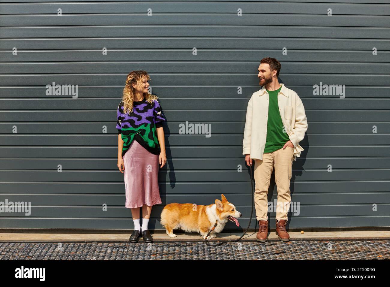 coppia felice ed elegante che cammina con il cane corgi vicino alla porta grigia del garage, compagni di animali Foto Stock