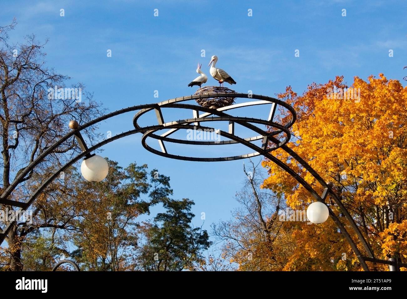 Parco cittadino in autunno. Arco in metallo con lanterne di illuminazione. In cima all'arco - una scultura a forma di nido e due cicogne. L'arco contro Foto Stock