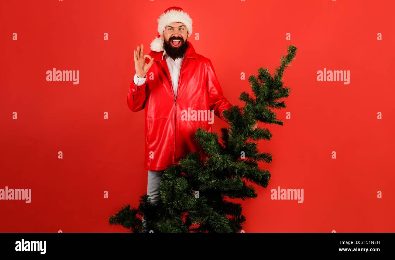 Felice uomo barbuto nel cappello di Babbo Natale con l'albero di Natale che mostra un cartello ok. L'uomo di Babbo Natale in giacca di pelle rossa con abete mostra un gesto discreto. Natale e.. Foto Stock