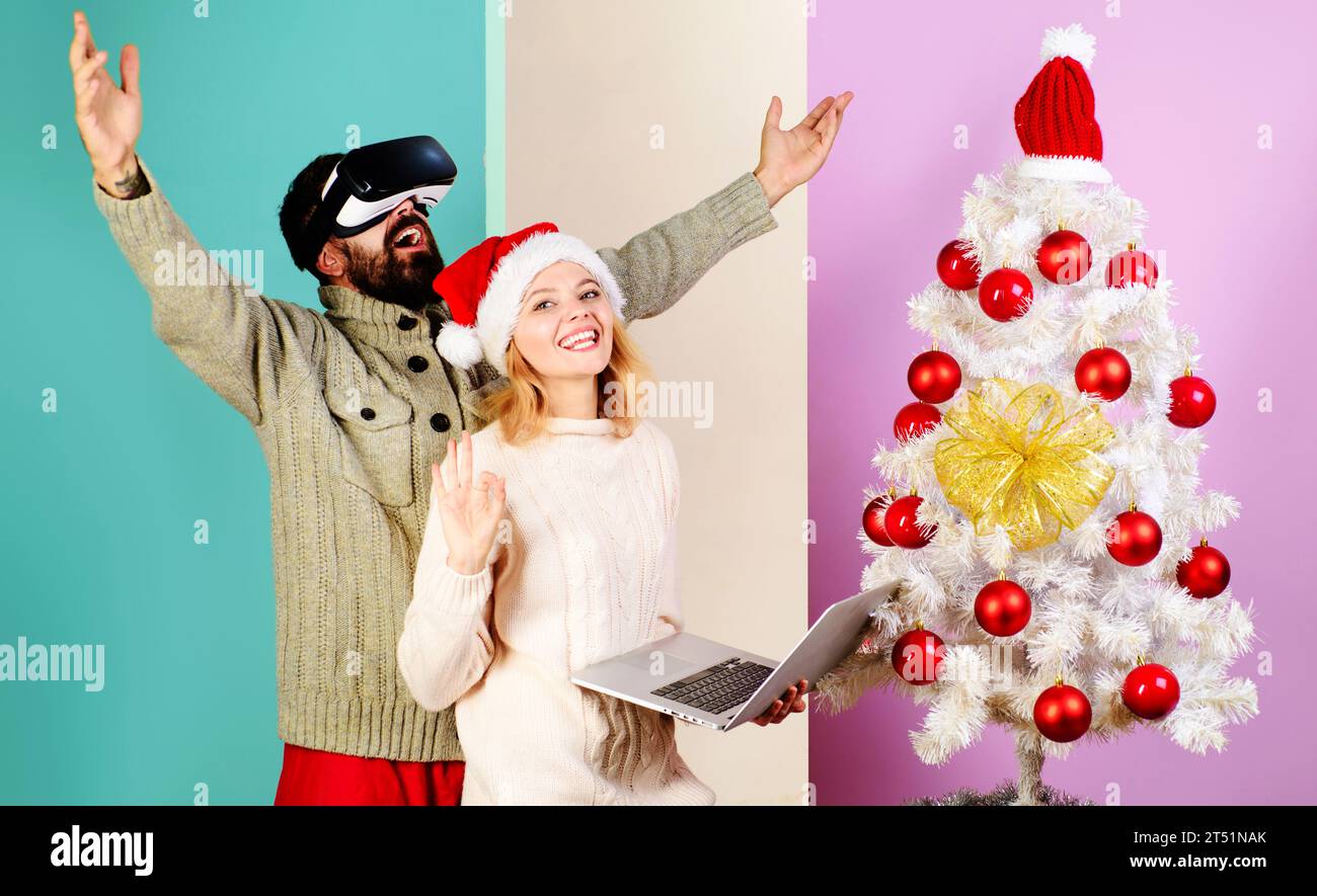 Famiglia felice vicino all'albero di Natale. Uomo barbuto con occhiali per realtà virtuale e donna sorridente con cappello di Babbo Natale con un cartello con lo spettacolo del notebook ok. tecnologia 3d. Coppia di Natale Foto Stock