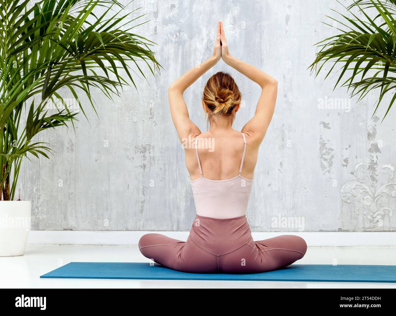 Vista posteriore di una donna che pratica la posa yoga di Sukhasana seduta a gambe incrociate sul pavimento con le mani sollevate per il relax, la meditazione e l'allineamento del corpo Foto Stock